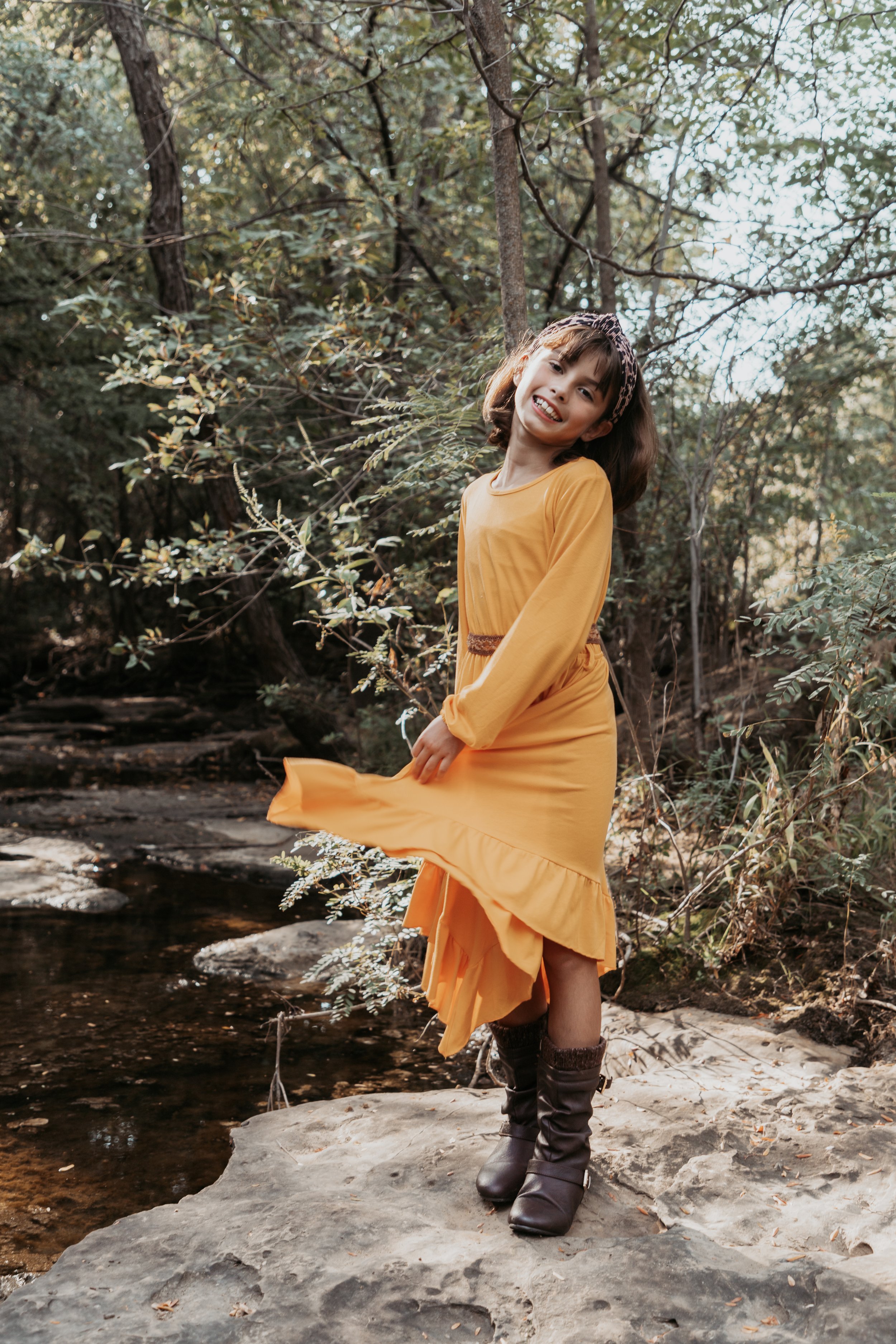 A woman wearing a yellow dress with brown boots stands on a rock near a creek in a wooded area, smiling and looking at the camera.