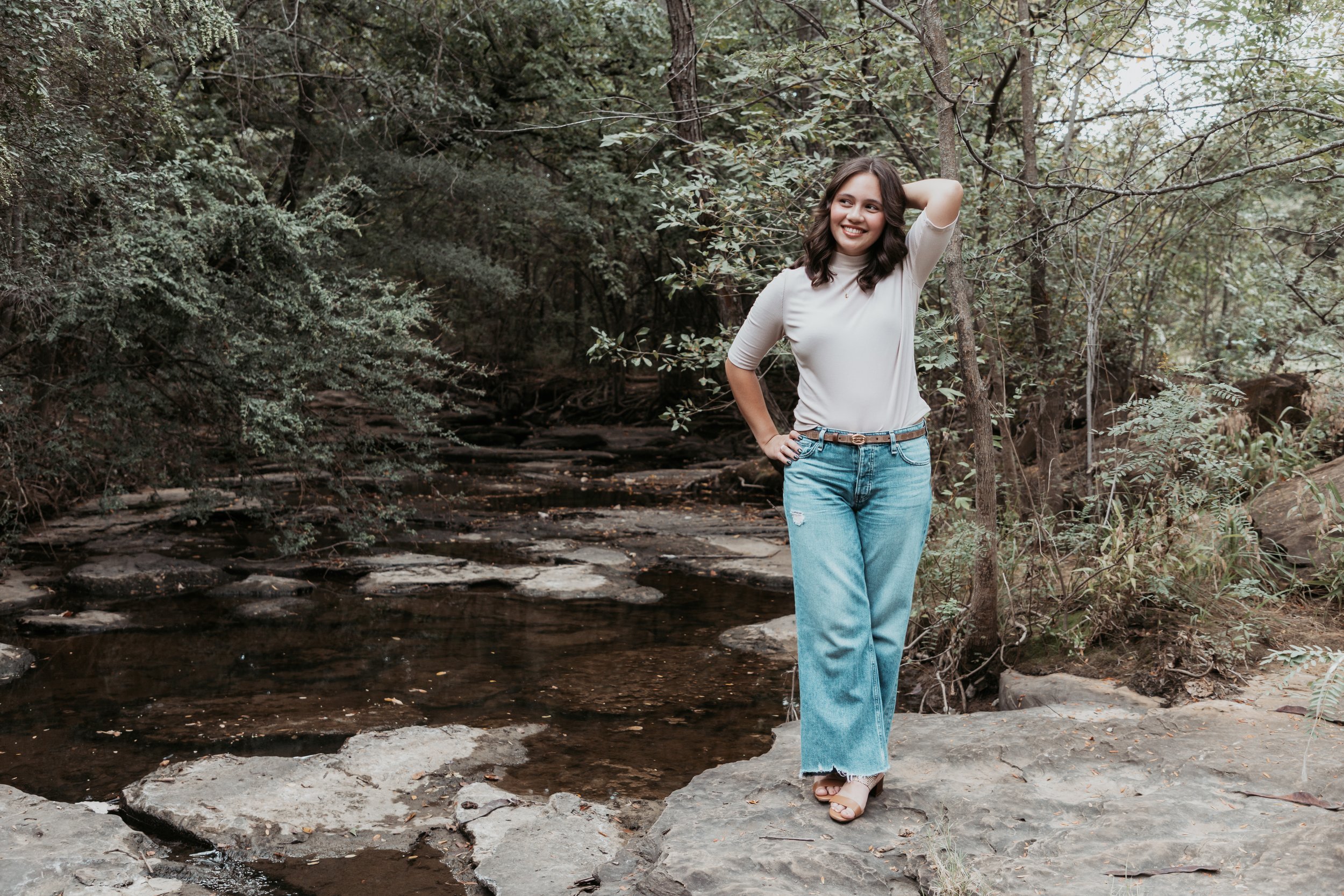 A woman with shoulder-length dark hair, wearing a white blouse and blue jeans, standing on a rock near a small creek in a forested area, smiling and looking to the side.