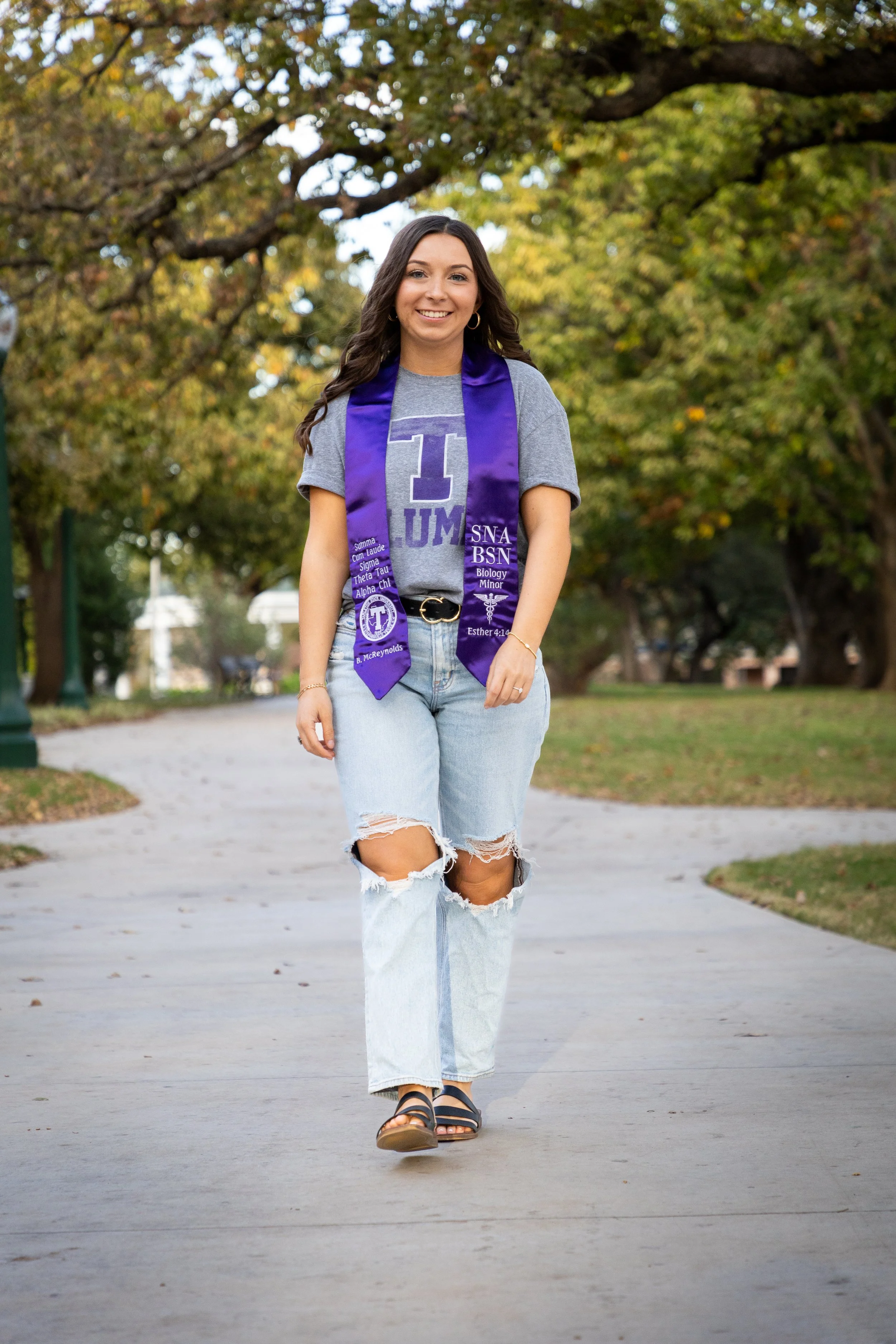 Young woman walking on a park path wearing a gray T-shirt, faded ripped jeans, sandals, and a purple graduation stole, smiling, autumn trees in the background.