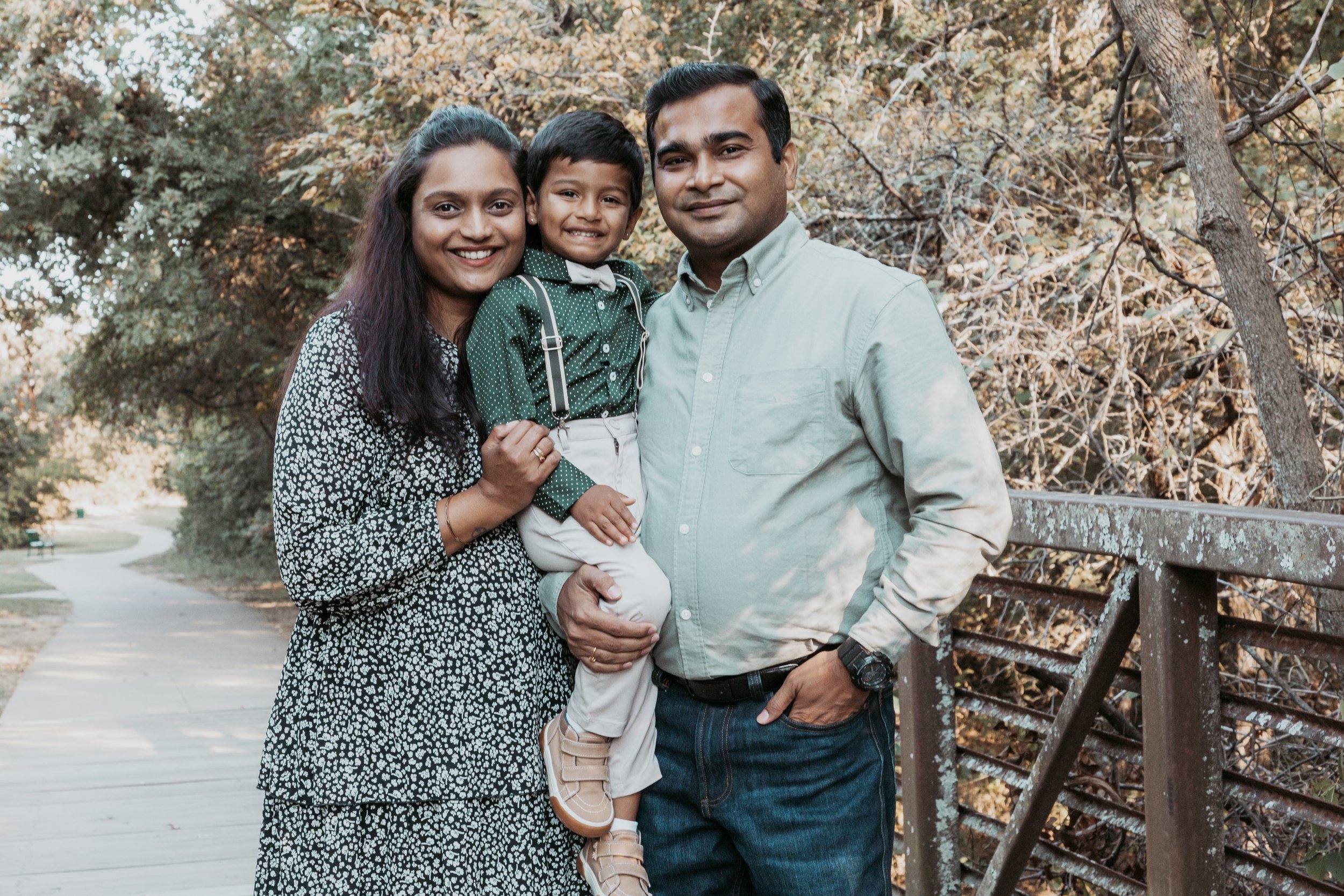 A happy family of three stands together outdoors on a walking trail, surrounded by trees with autumn leaves. The mother is wearing a black and white patterned dress, the father is wearing a light green shirt and jeans, and their young son is dressed 