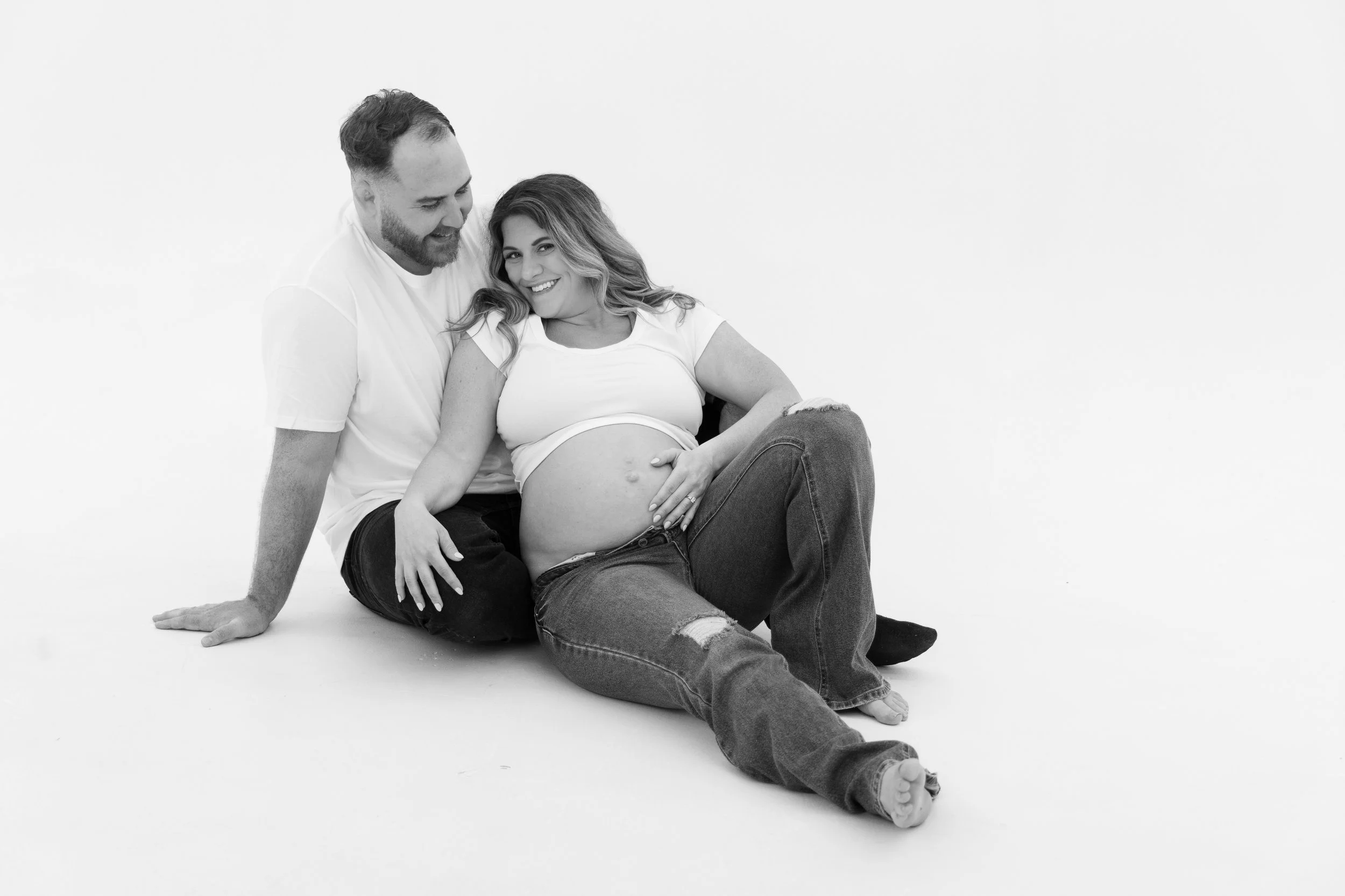 A pregnant woman with long wavy hair smiling while sitting on the floor, gently touching her belly, with a man sitting behind her, smiling and looking at her, both in front of a plain white background.