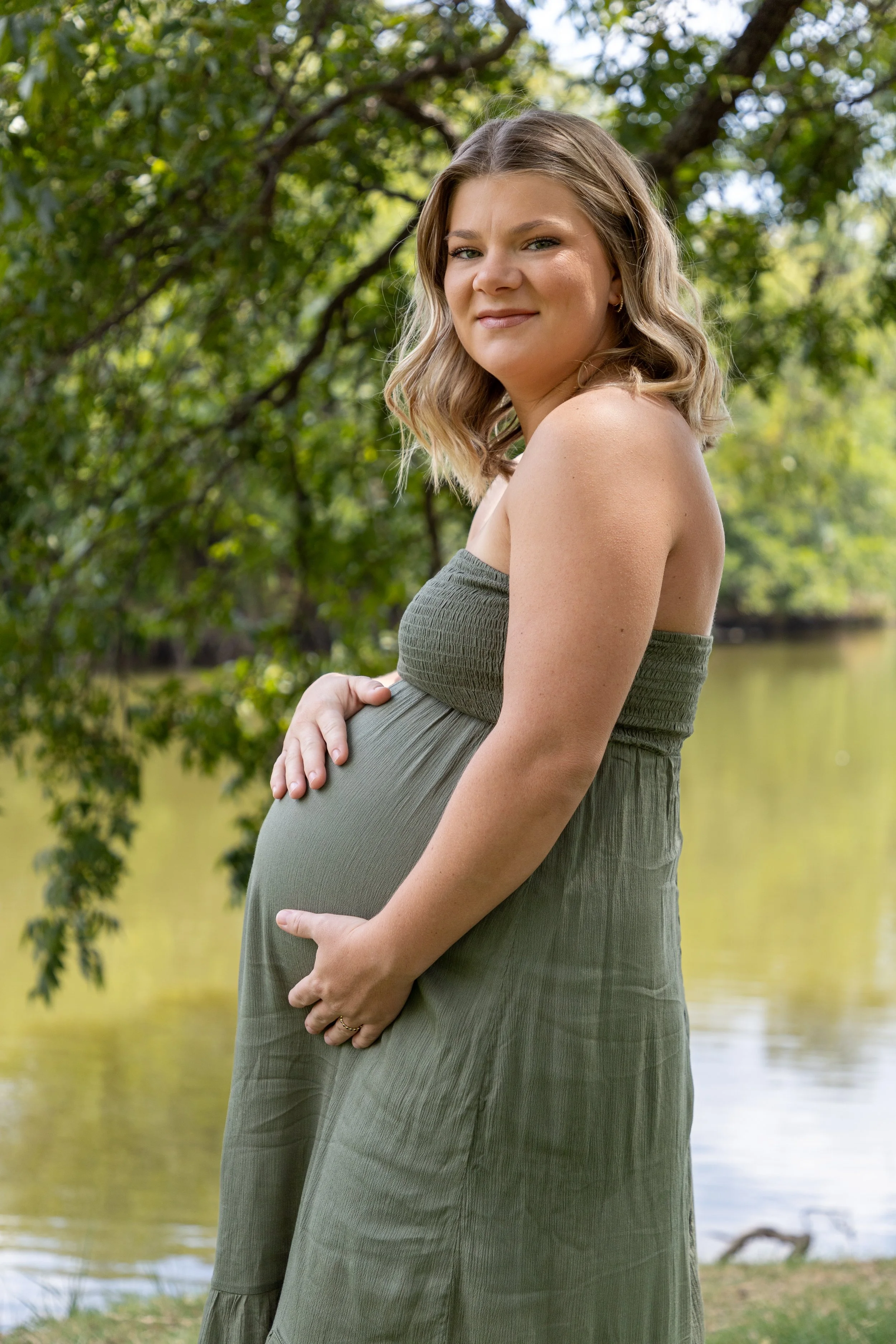 Pregnant woman standing outdoors near a lake, holding her belly with one hand, smiling gently, and wearing a strapless green dress, surrounded by trees and greenery.