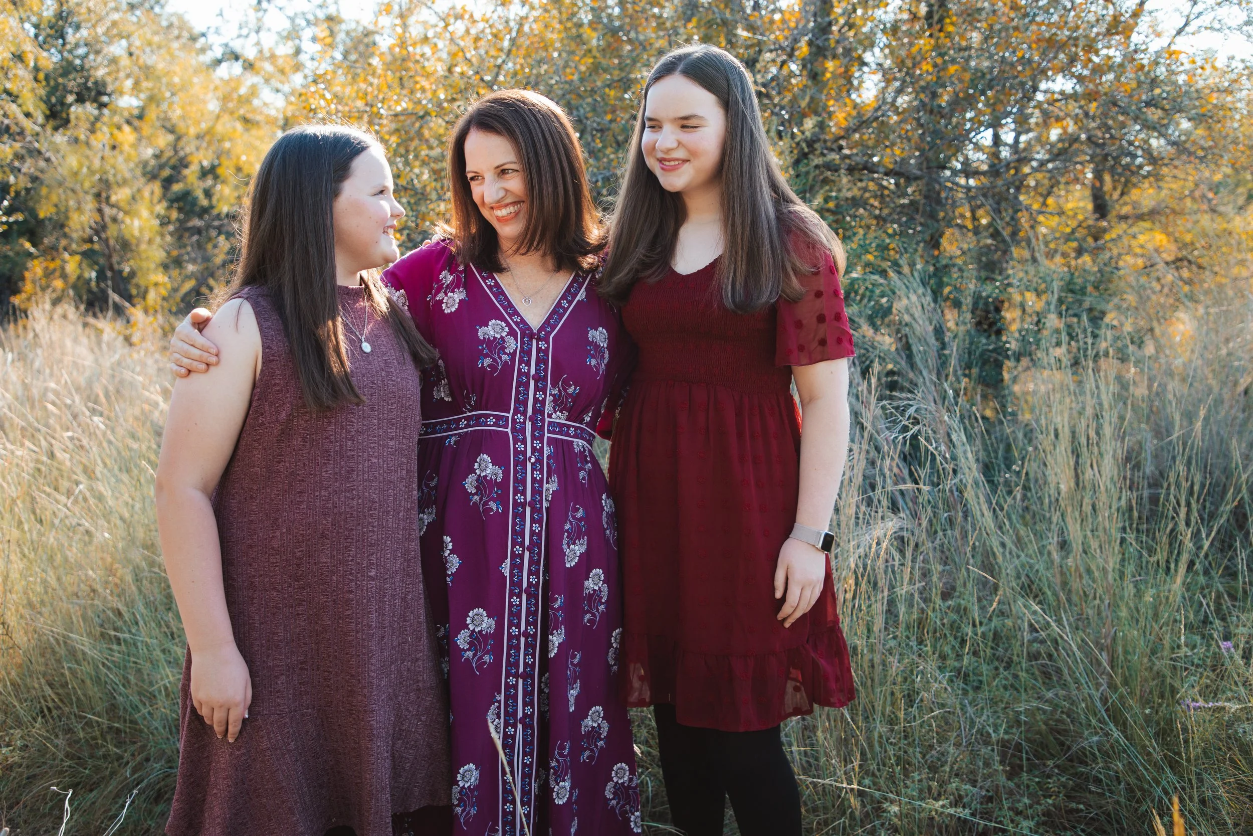 Three women standing outdoors in a field of tall grass with autumn trees in the background, smiling and looking at each other.