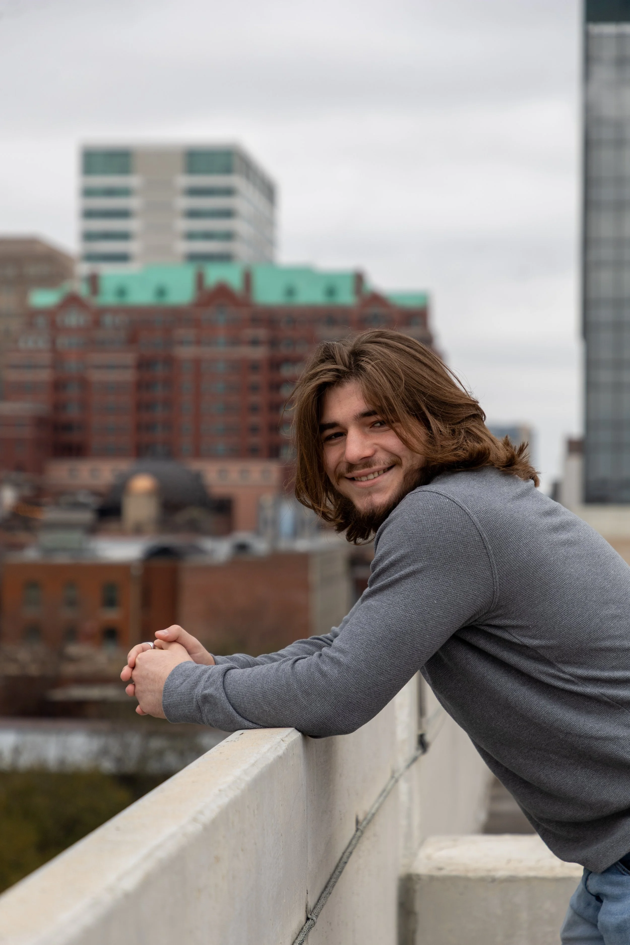 A young man with long hair, smiling, wearing a gray long-sleeve shirt, leaning on a concrete railing on a rooftop with a cityscape in the background.