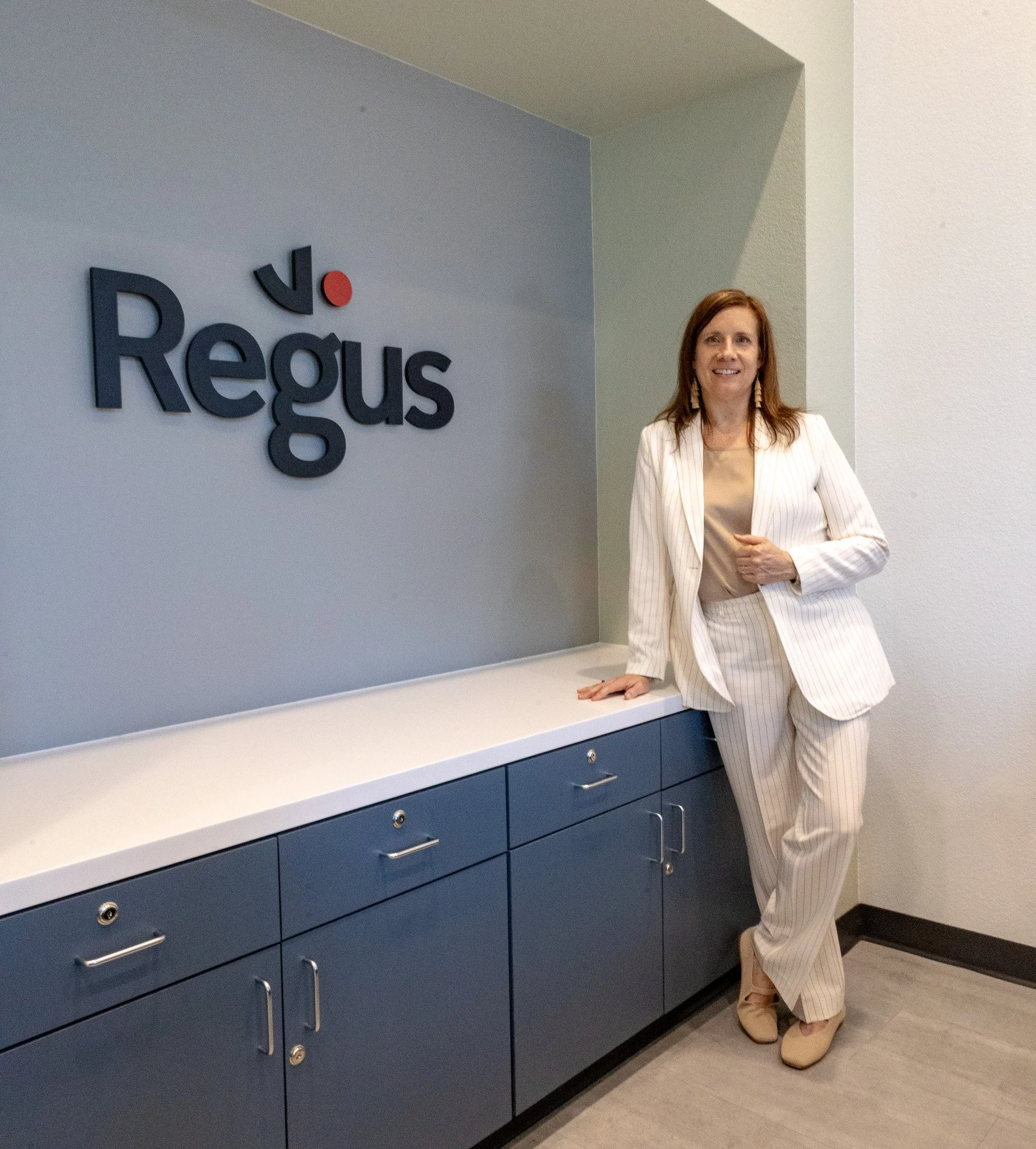 A woman in a cream-colored pinstripe suit standing next to a blue cabinet with a logo on the wall that reads 'Regus' in a modern font.