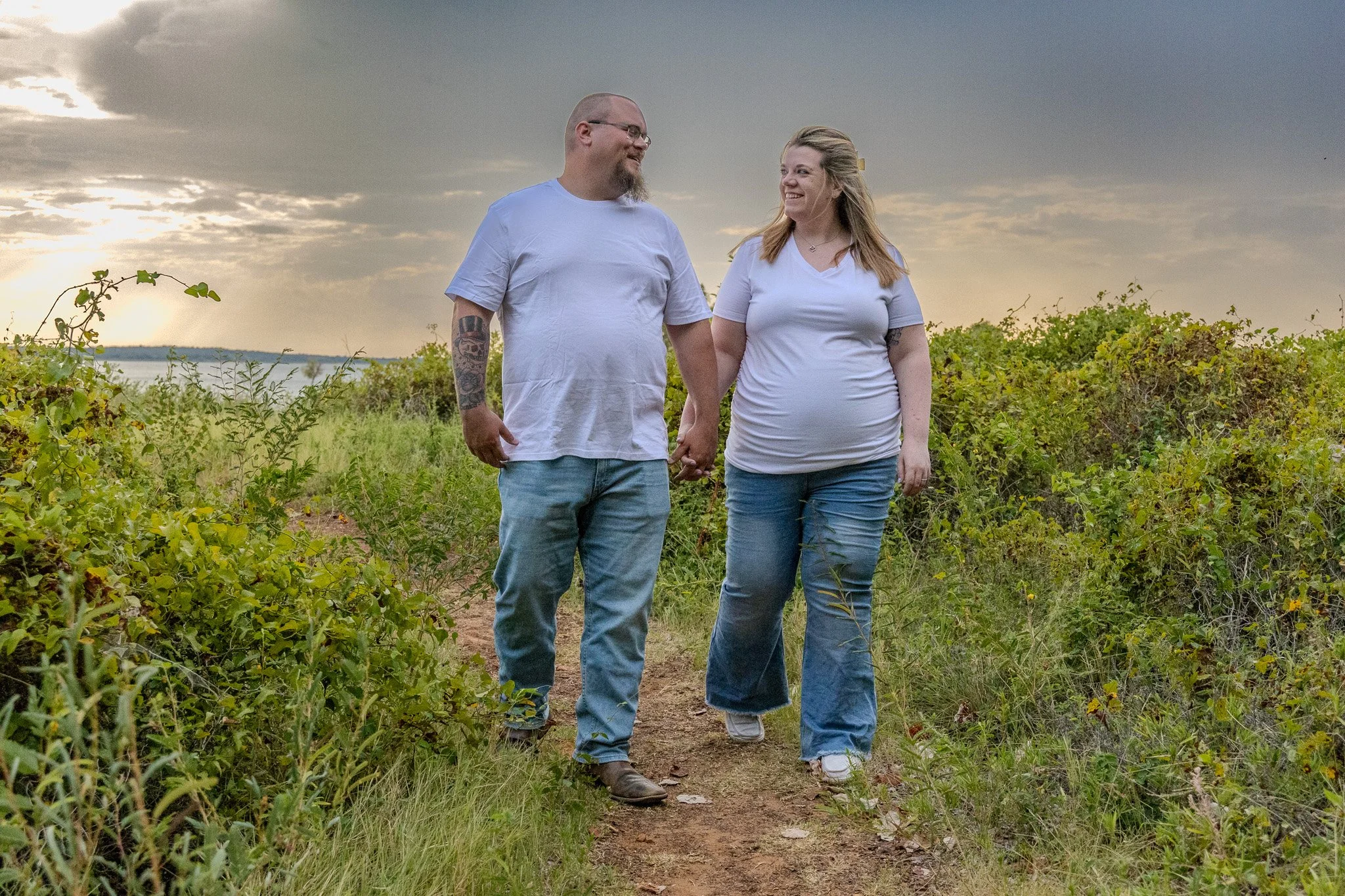A man and woman holding hands walking along a dirt path through greenery, with a body of water and a cloudy sky in the background.