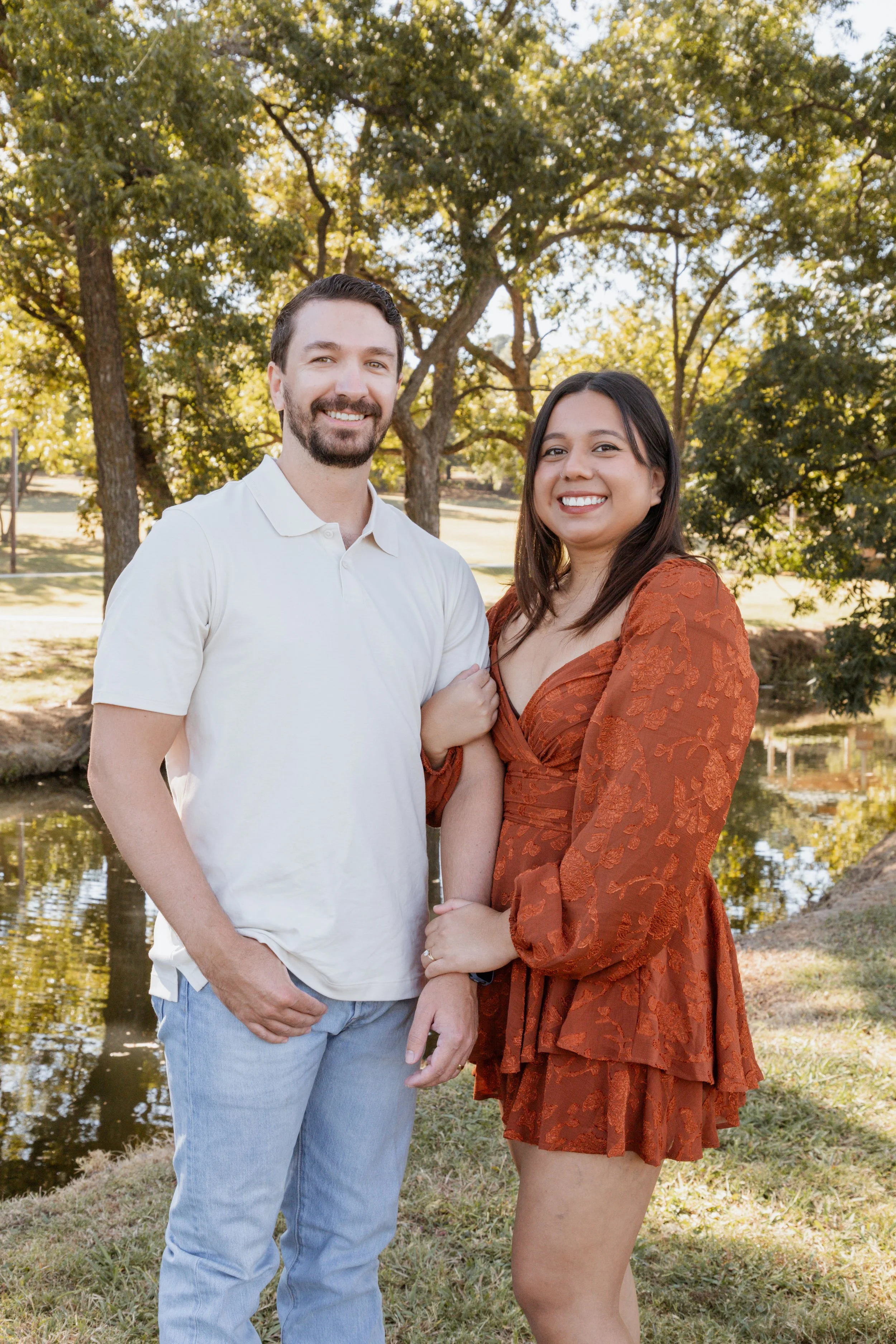 A smiling couple standing outdoors near a pond during daytime, surrounded by trees with green leaves, with the woman wearing a rust-colored dress and the man in a white polo shirt and light jeans.