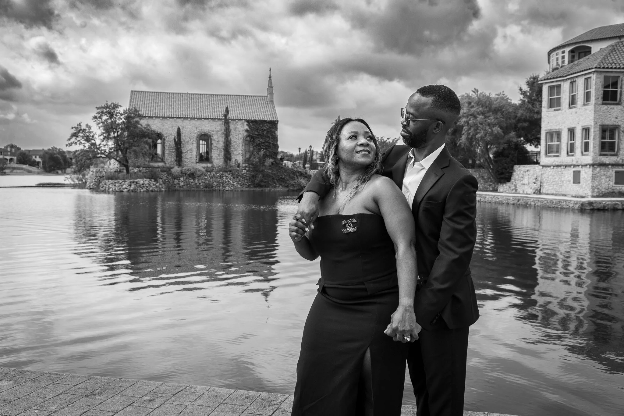 A man and woman hold hands and pose together near a body of water with houses and a church in the background on a cloudy day.
