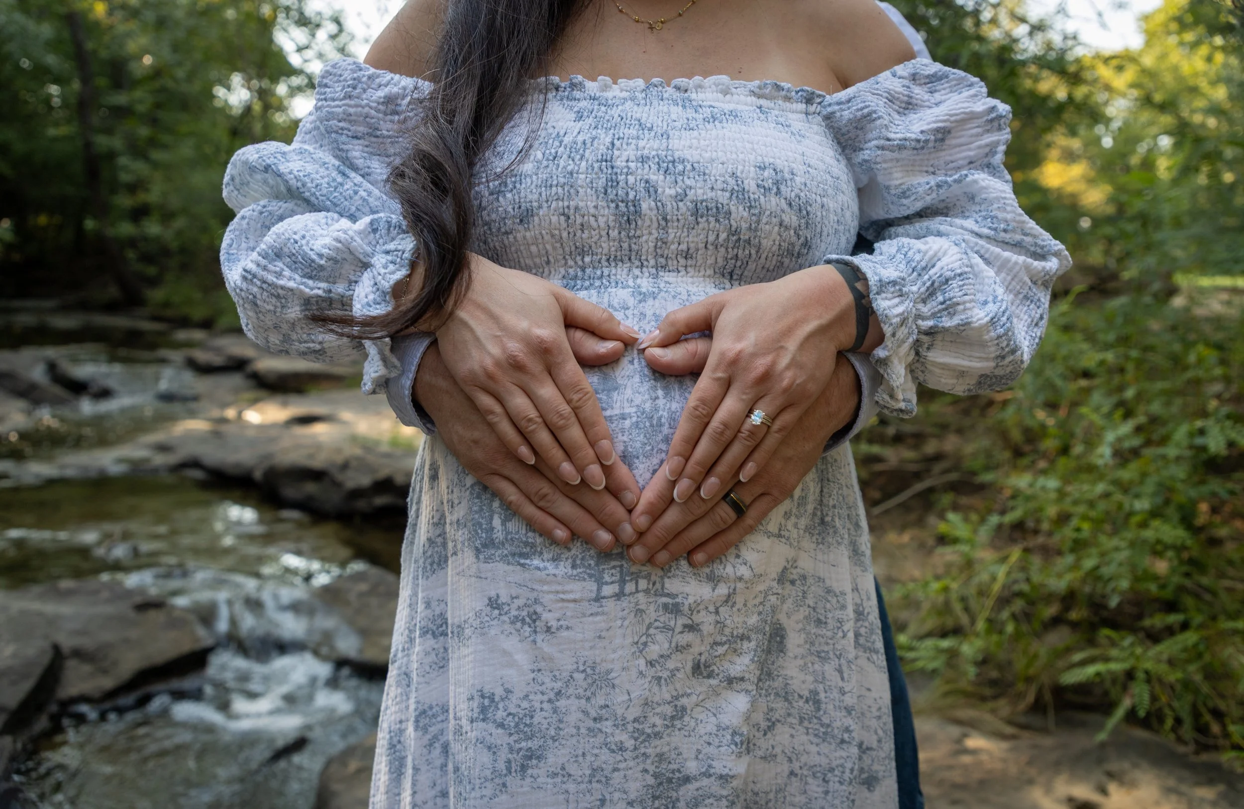 A woman in a light-colored off-shoulder dress forming a heart shape with her hands on her stomach in a natural outdoor setting by a creek.