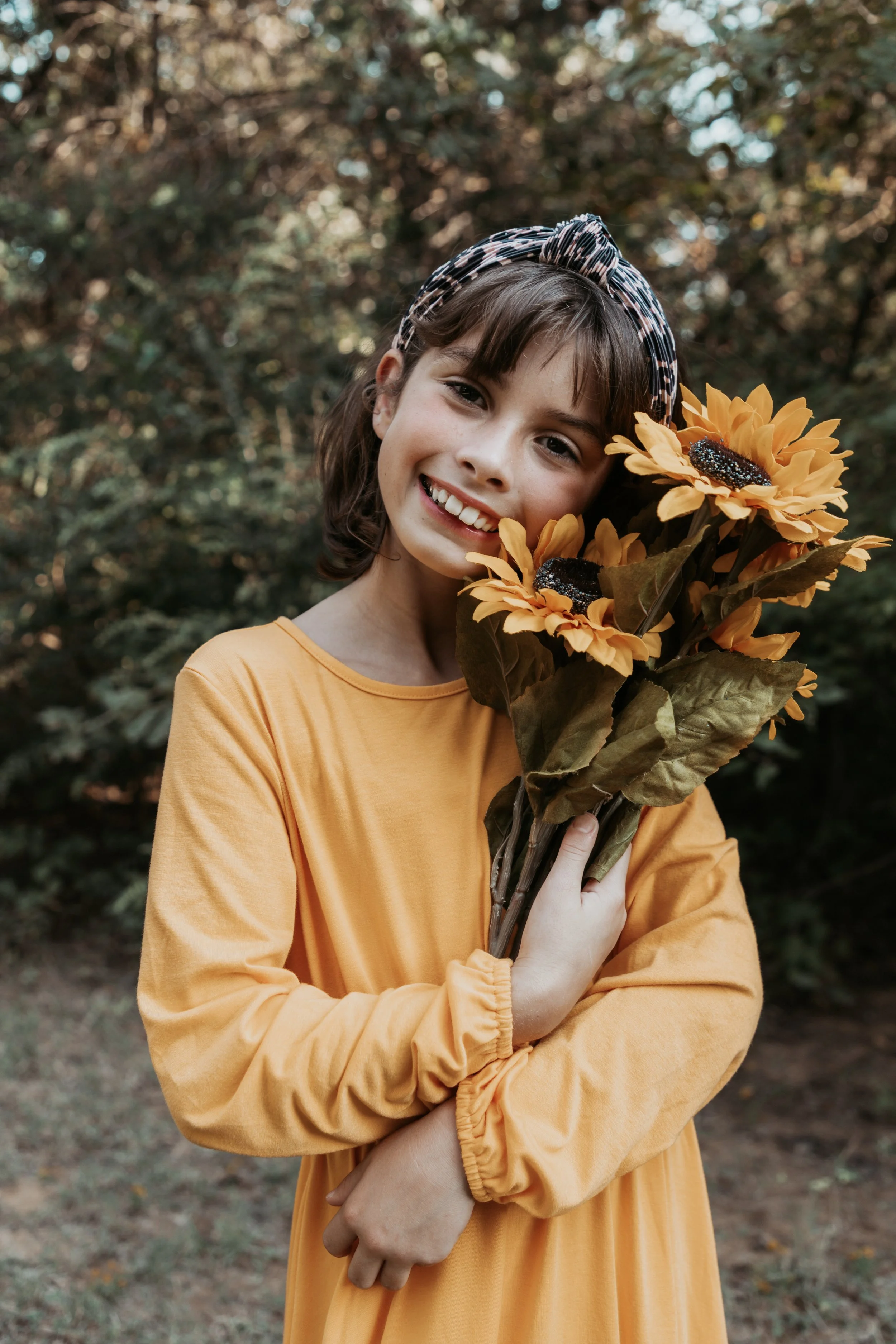 A young girl wearing an orange dress and a patterned headband is holding a bouquet of artificial sunflowers, smiling at the camera outdoors with a blurred natural background.