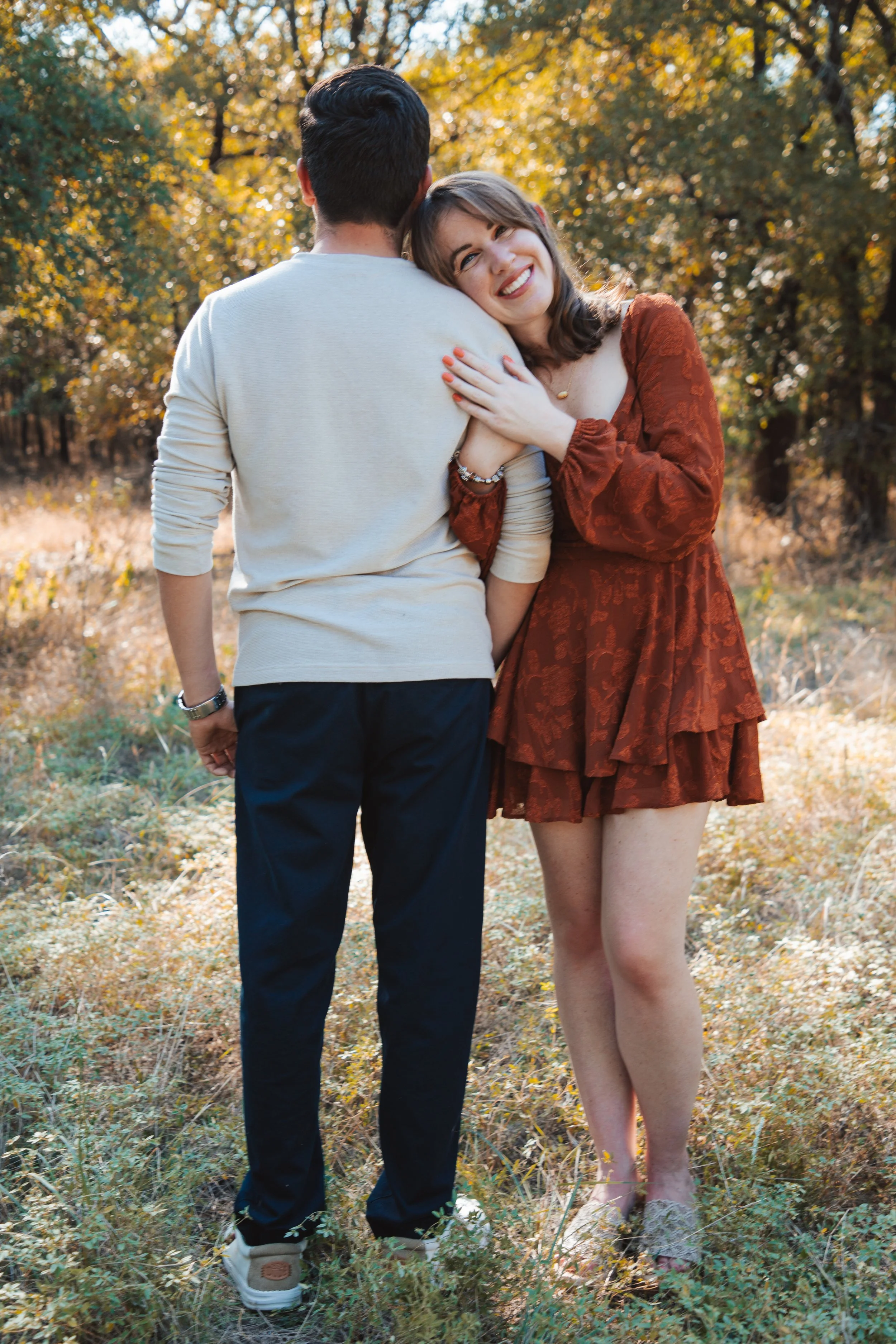A young woman in a rust-colored dress smiling and hugging a man in a light sweater outdoors during autumn.