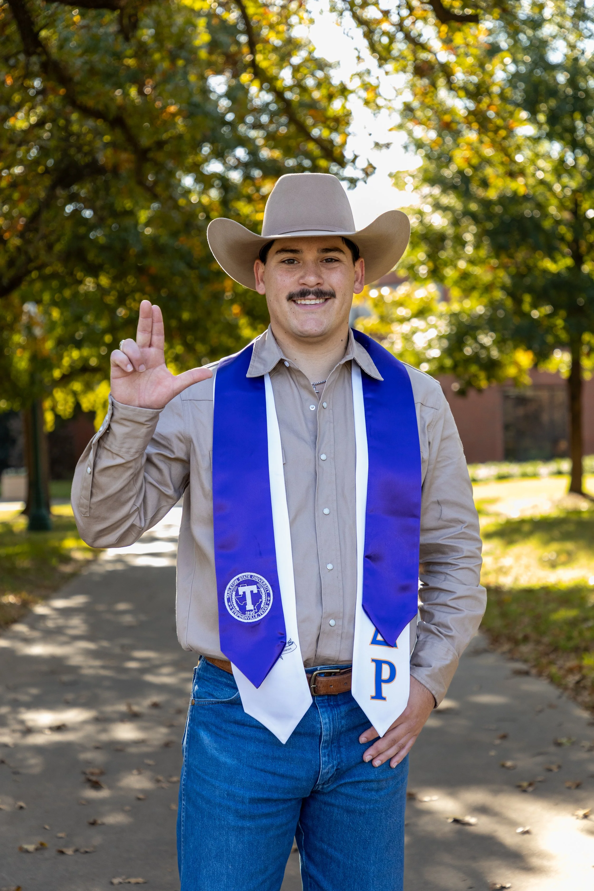 A young man wearing a cowboy hat, beige button-up shirt, and blue jeans, standing outdoors on a park path with trees in the background. He is smiling and holding hand in the Greek sign. He has a blue and white sash with university logos and Greek let