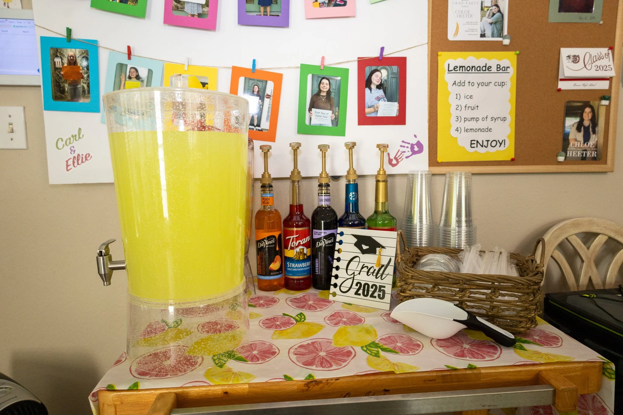A lemonade bar setup with a large container of lemonade, bottles of flavored syrup, plastic cups, and a sign that says "Grad 2025". There are colorful photos hanging on a bulletin board in the background.