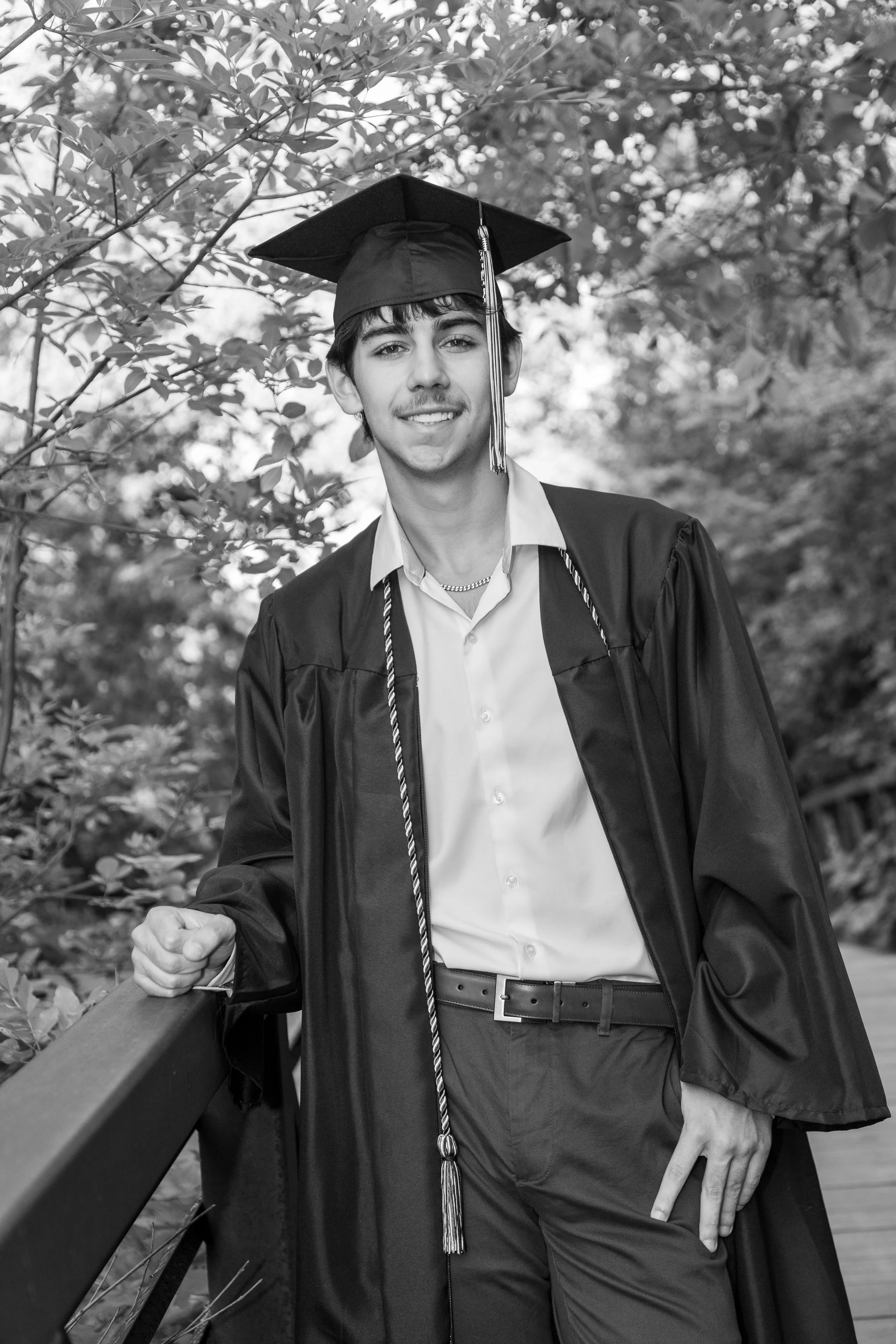 Young man in graduation cap and gown smiling outdoors on a wooden bridge with trees in the background.