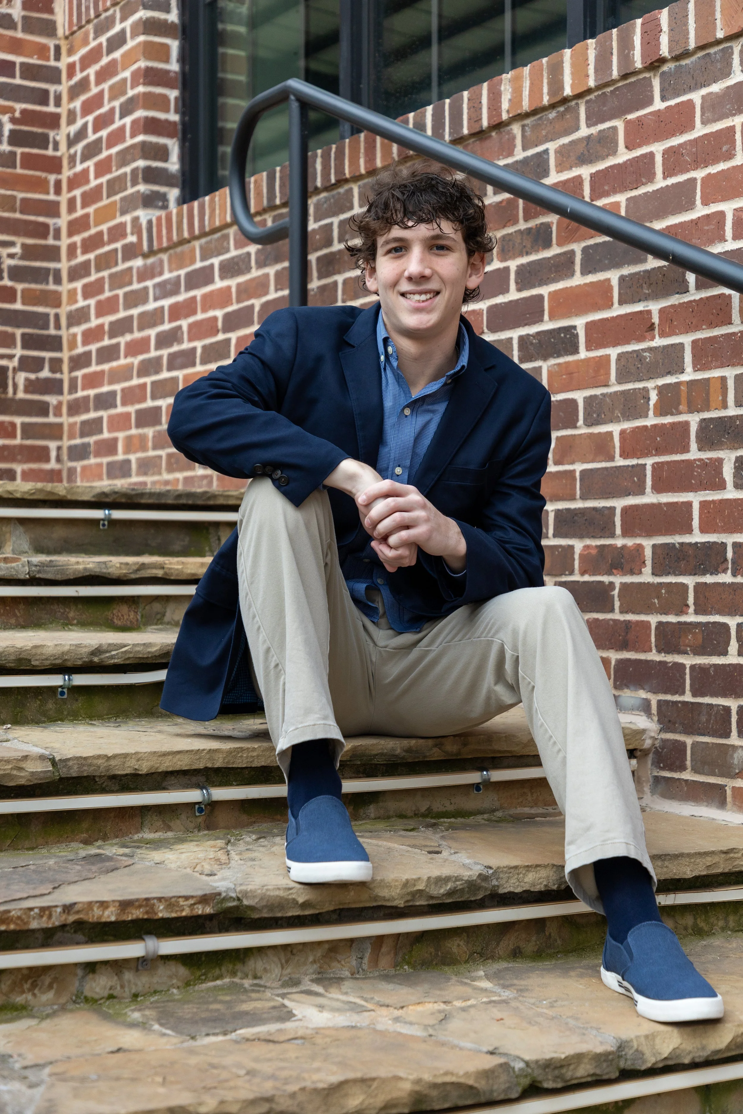 A young man with curly hair, wearing a navy blazer, blue shirt, khaki pants, and navy slip-on shoes, sitting on outdoor stone stairs in front of a brick building and smiling at the camera.