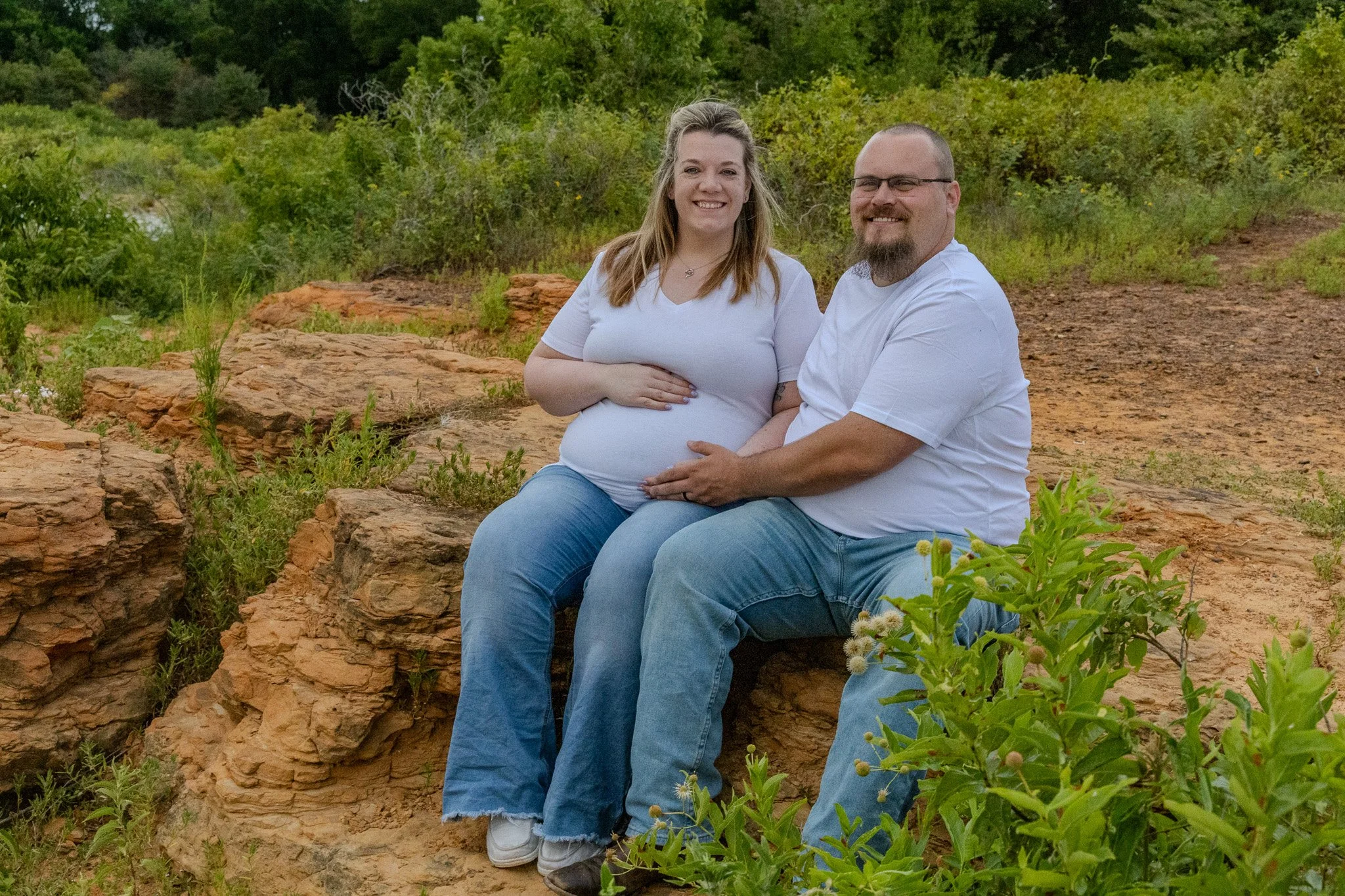 A pregnant woman and a man sitting on a large rock in a natural outdoor setting with greenery and bushes, smiling at the camera.