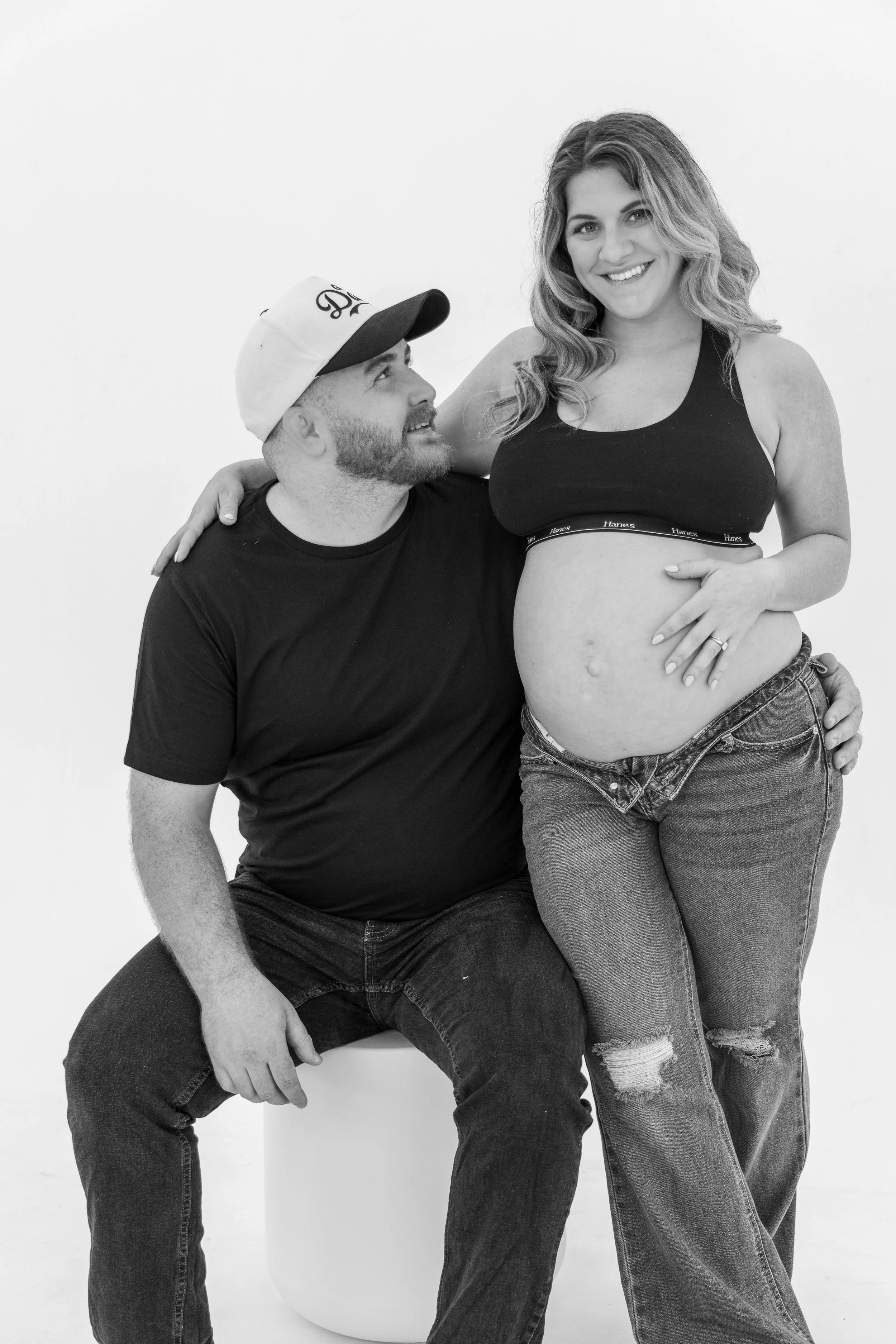 A smiling pregnant woman with long, wavy hair and a sleeveless black sports bra, standing and touching her pregnant belly, while her partner, a man with a beard wearing a baseball cap and a black t-shirt, is sitting with his hand on her leg, looking 