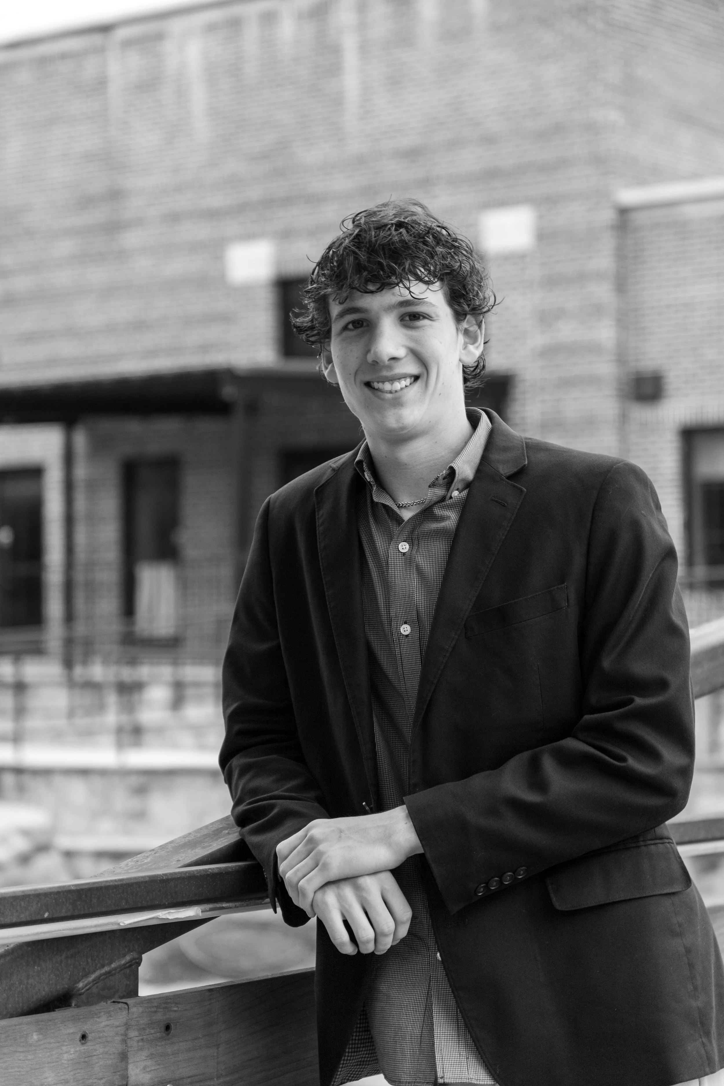 Black and white photo of a young man wearing a blazer and button-up shirt, smiling, standing outdoors near a wooden railing with a brick building in the background.