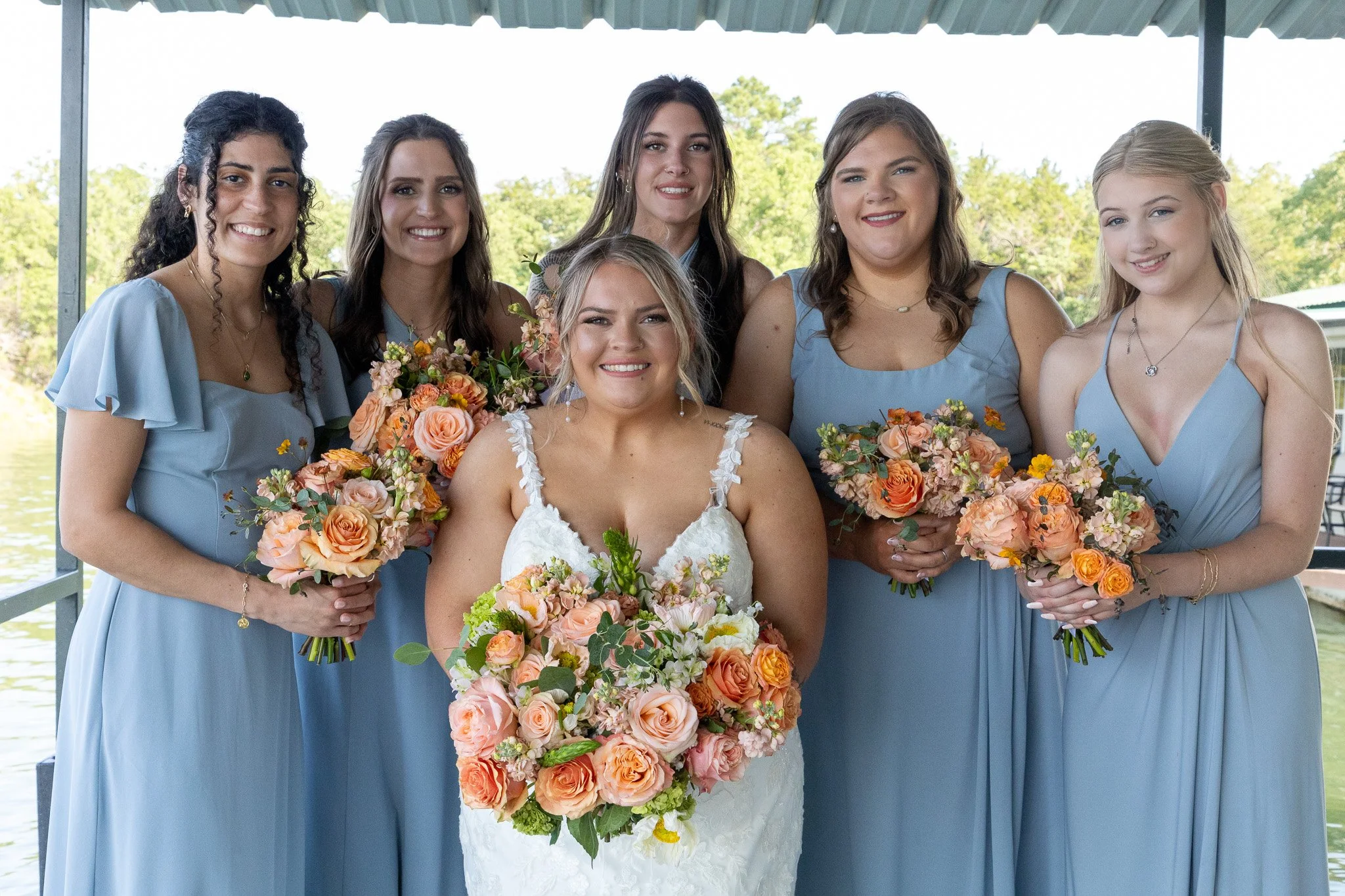 Bride in white dress smiling, surrounded by six bridesmaids in light blue dresses holding bouquets of peach, pink, and orange flowers, outdoors near water with a green background.