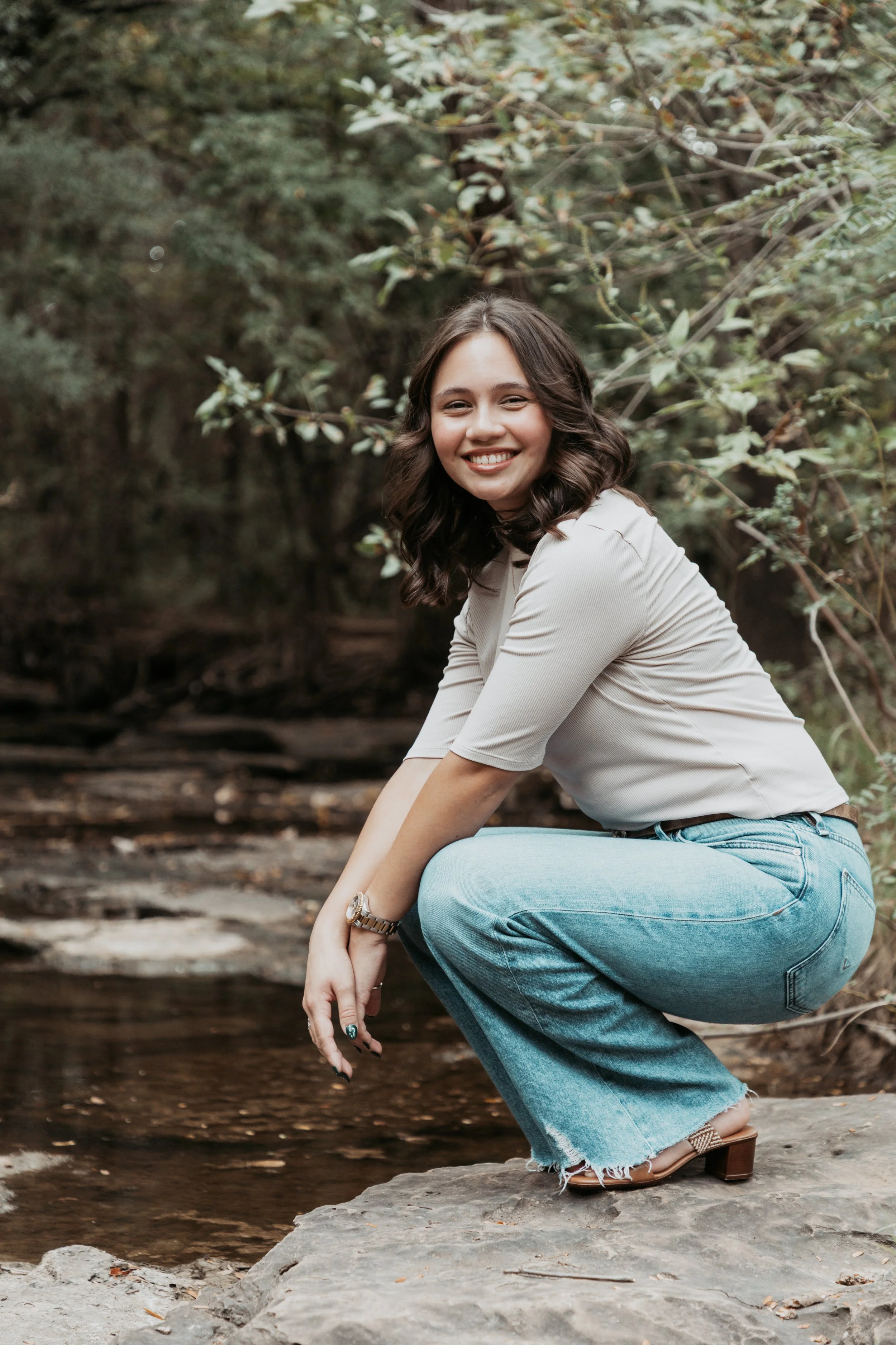 Young woman crouching on a rock by a creek in a wooded area, smiling at the camera.