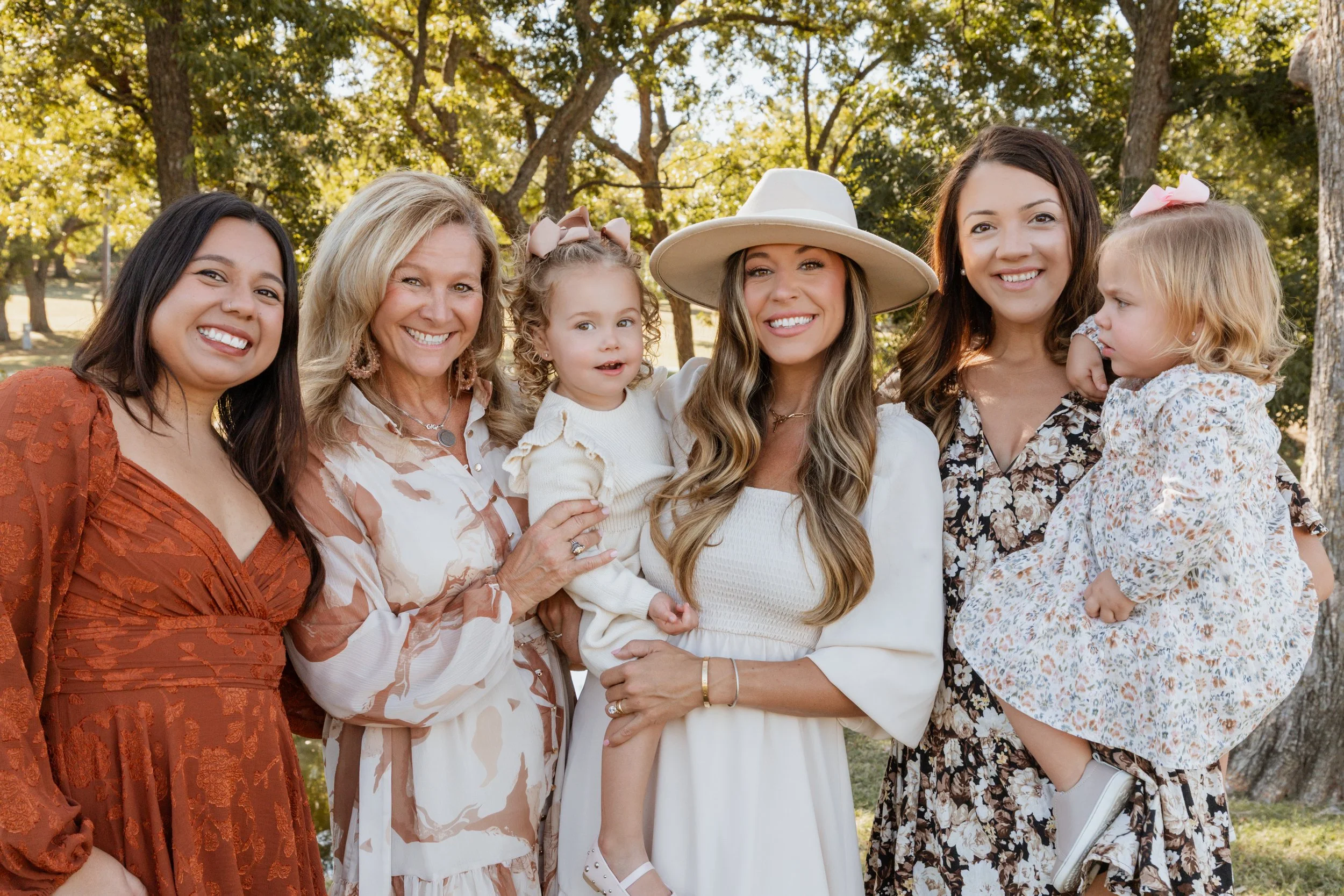 Group of six women and two young girls outdoors in a park, smiling for a photo on a sunny day.