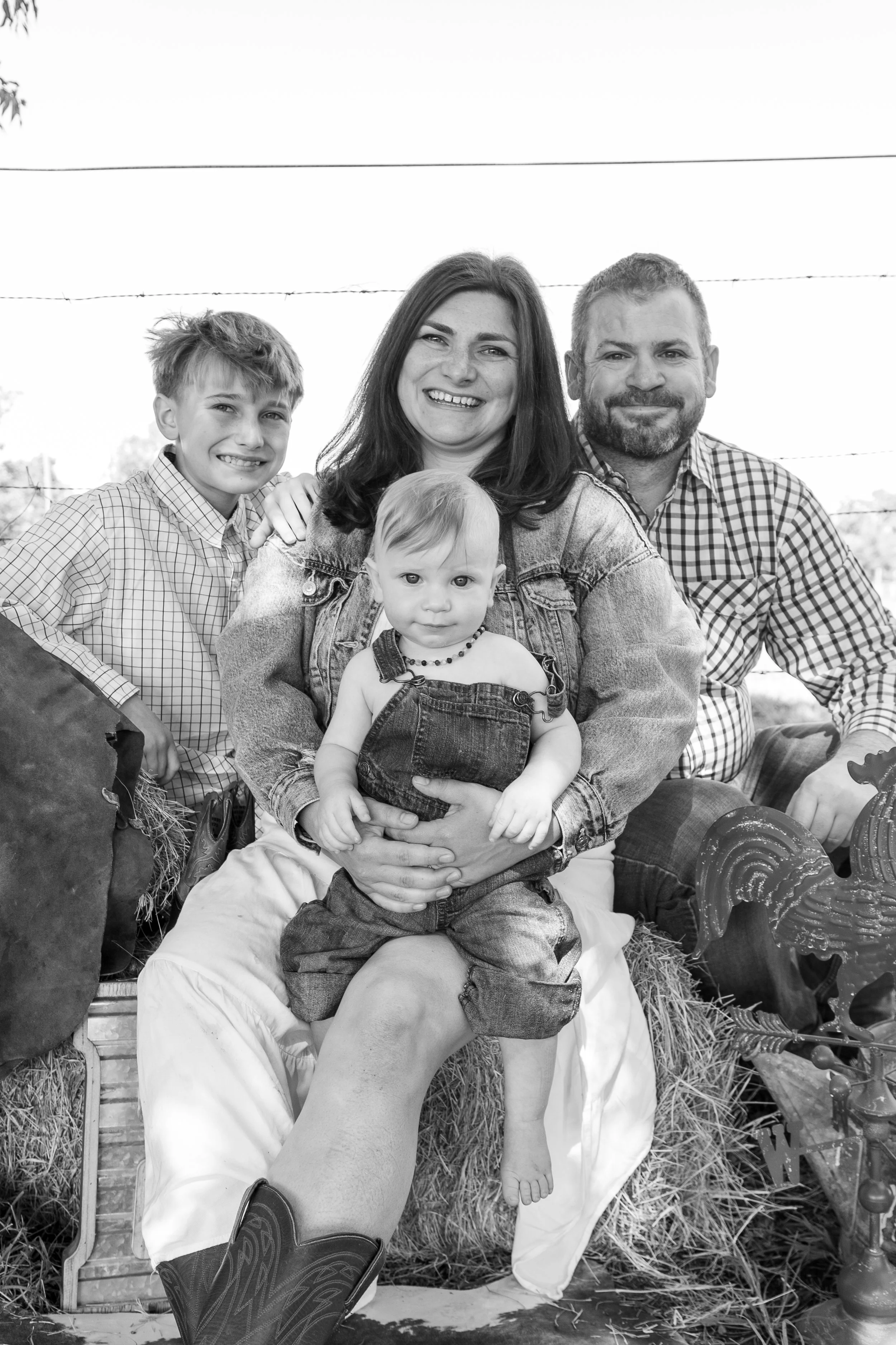 Black and white photo of a happy family of four outdoors, with a woman holding a toddler girl, a young boy standing beside them, and a man sitting behind them. They are sitting on hay.