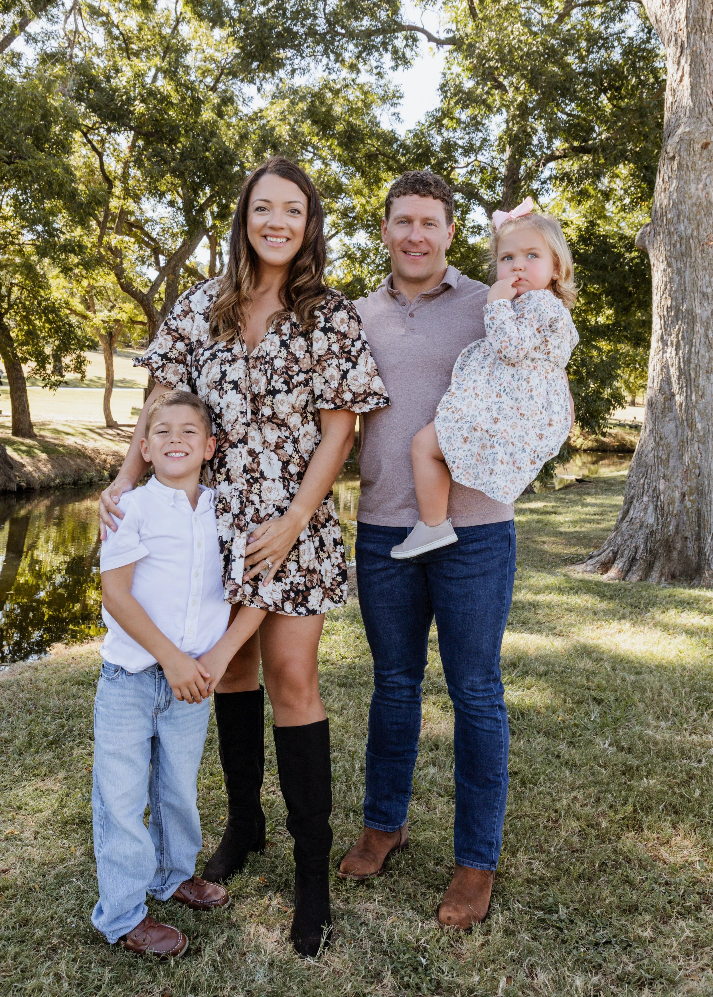 A family of two adults and three children standing outdoors near a pond and trees, smiling at the camera.