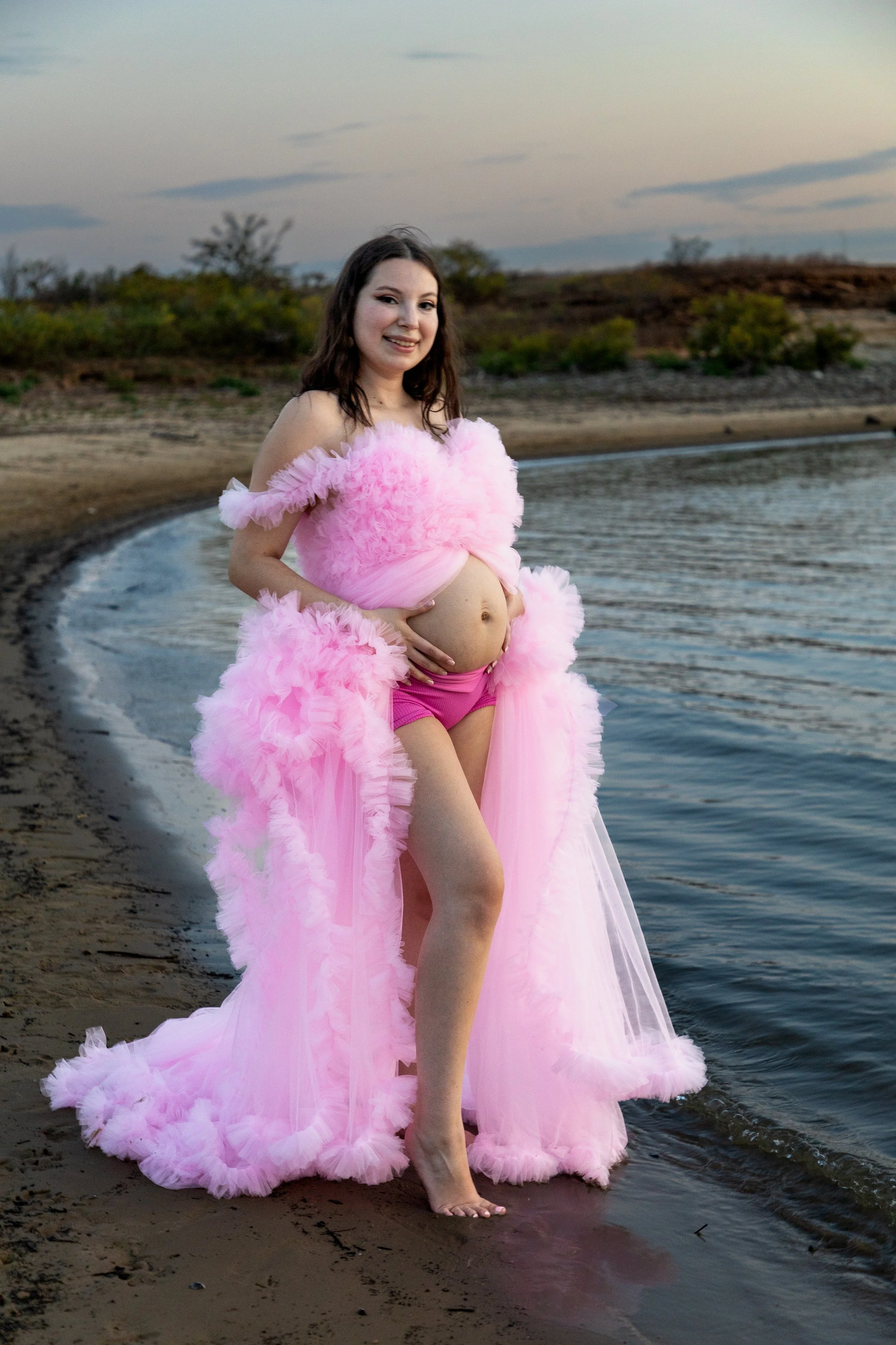 Pregnant woman standing on a beach at sunset, smiling, wearing a pink fluffy dress and shorts, with water and trees in the background.
