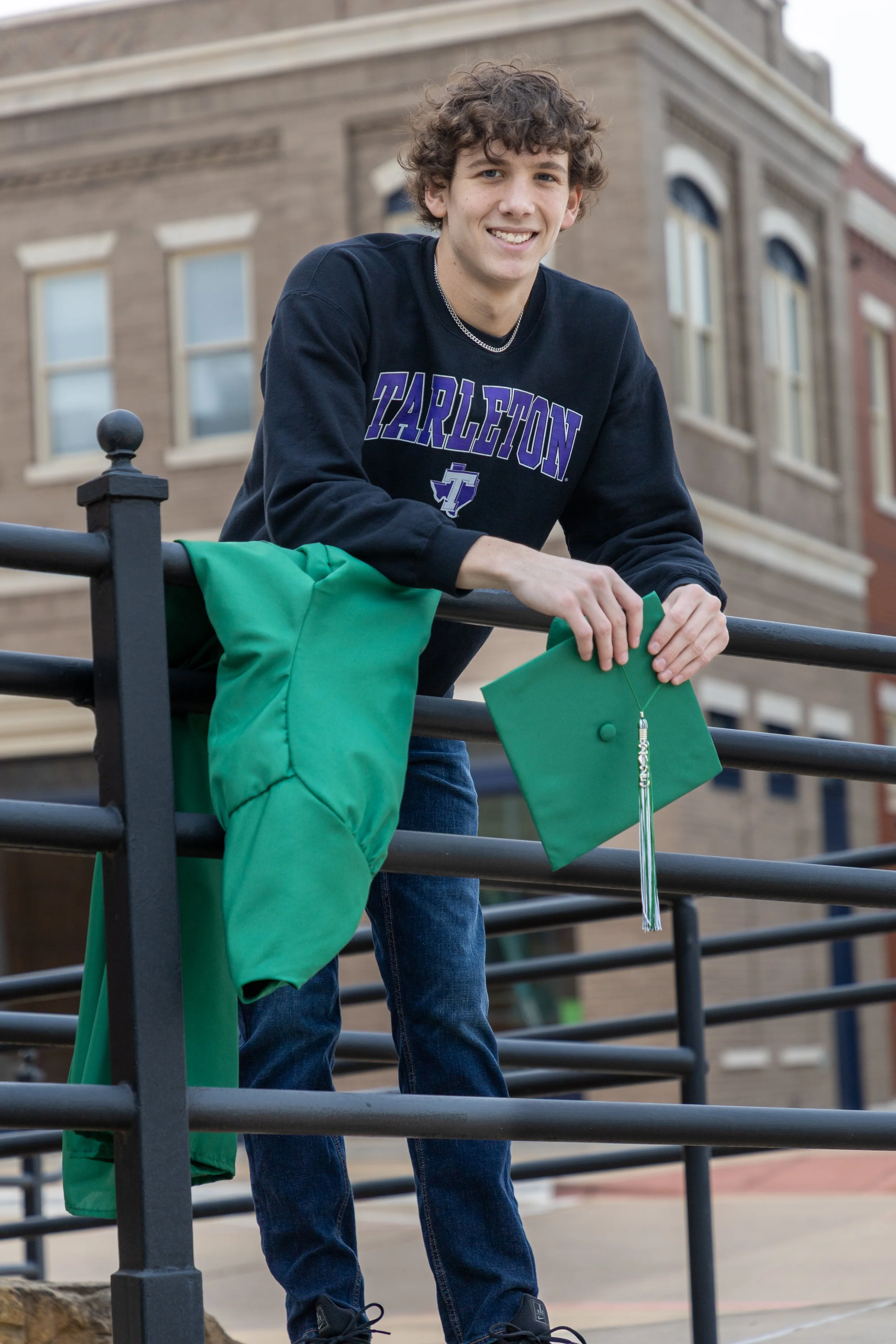 A young man with curly brown hair smiling, wearing a black sweatshirt with 'Tarleton' in purple, holding a green graduation cap and gown, standing on a railing outside in front of a brick building.
