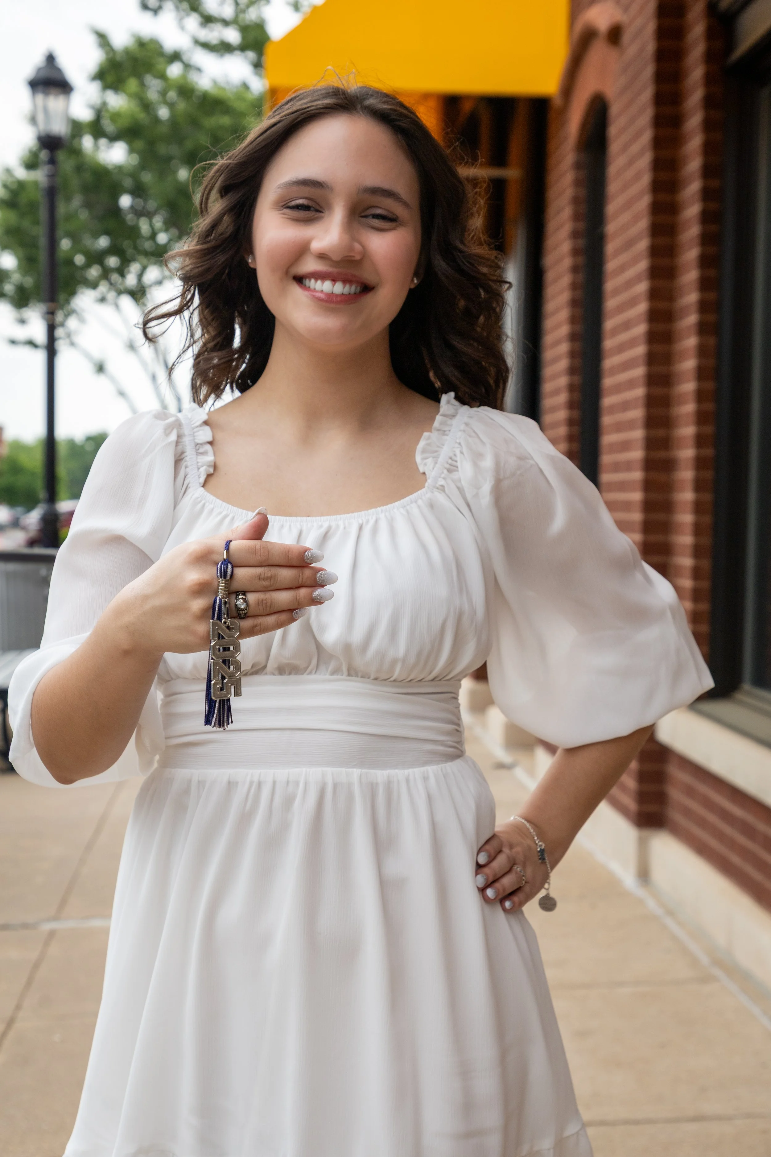 Woman in white dress holding keychain with '2023' charm, standing outside near brick building and trees