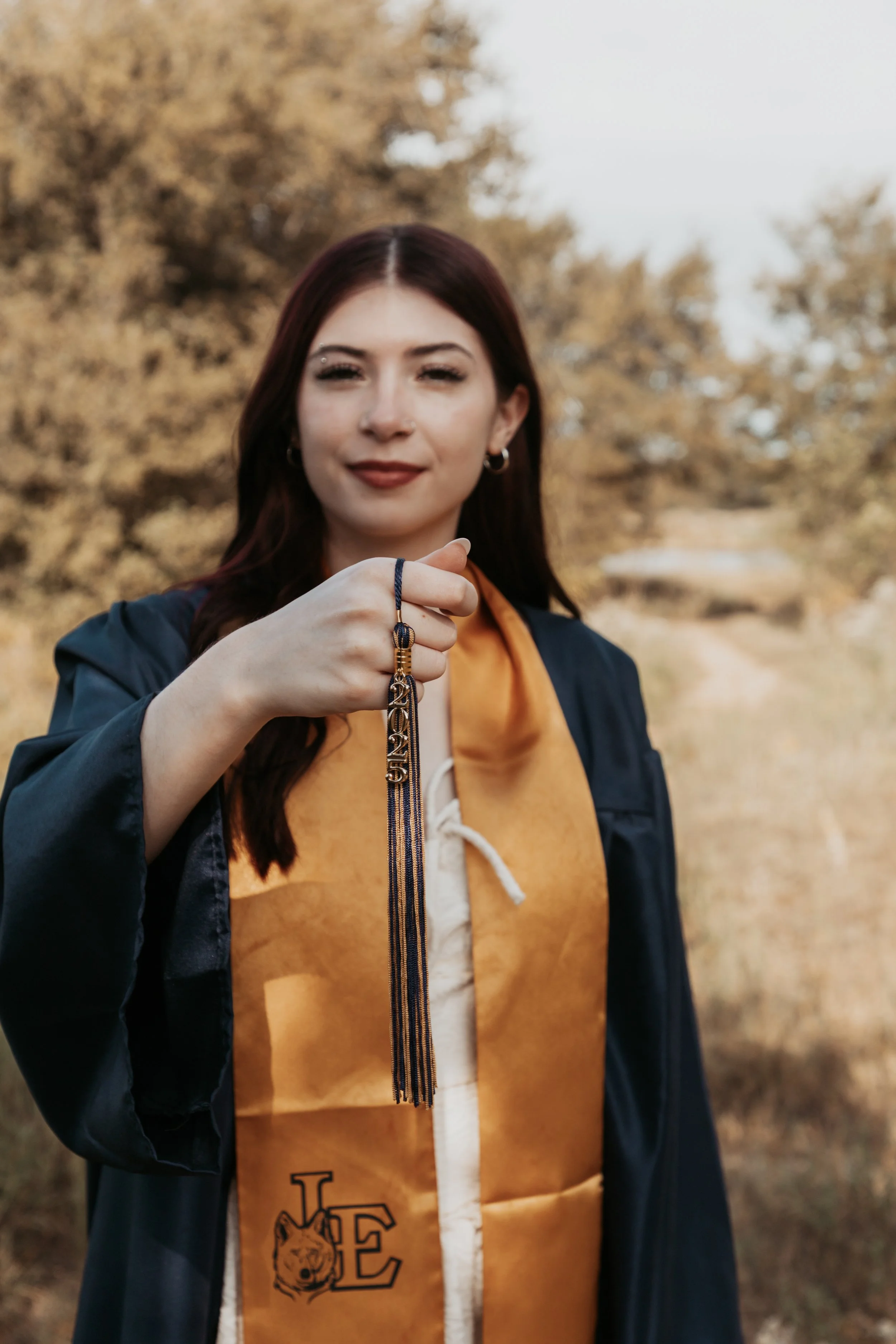 A woman wearing a graduation gown with a gold stole and holding a graduation tassel, standing outdoors with autumn trees in the background.