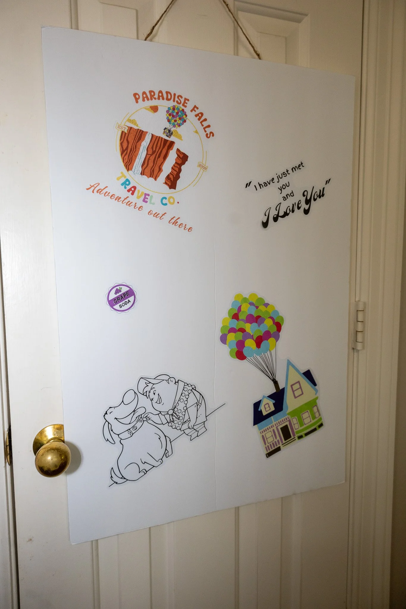Decorative wall poster with a colorful Paradise Falls logo from Up, a quote 'I have just met you and I Love You,' a drawing of characters Carl and Russell from Up, a small grape soda sticker, and an illustration of a house with balloons.
