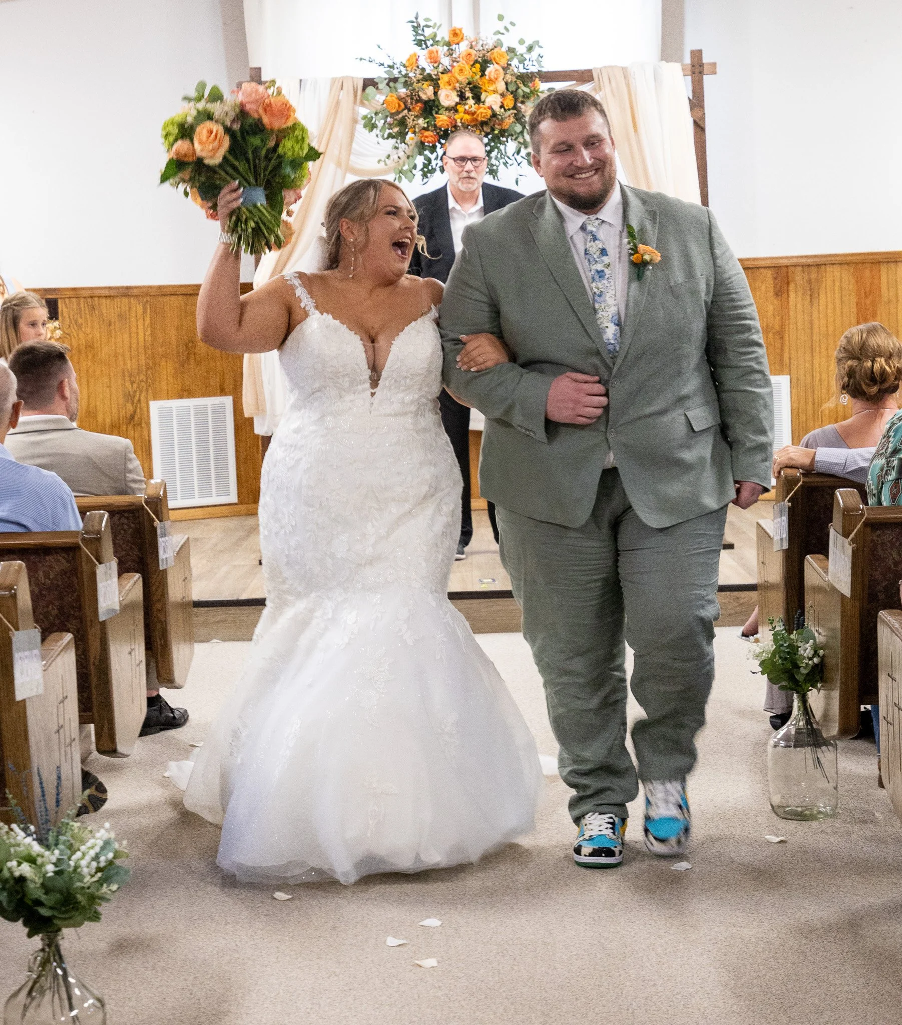 Bridal couple walking down the aisle during wedding ceremony, smiling with the bride holding a bouquet, officiant and guests in the background.