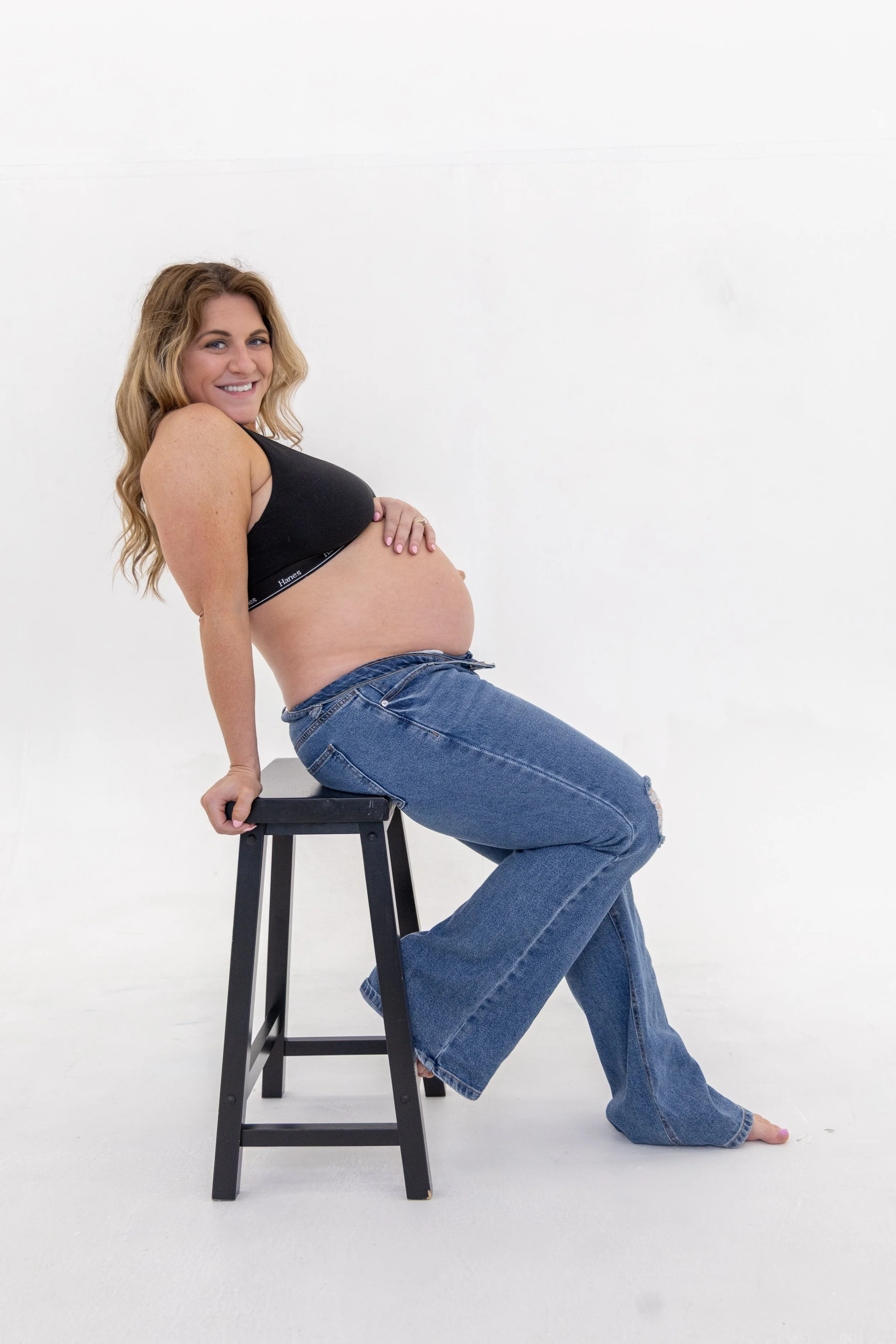 A smiling woman with long wavy blonde hair, wearing a black sports bra and blue jeans, posing in front of a white background while sitting on a black stool with her legs crossed and one foot resting on the floor.