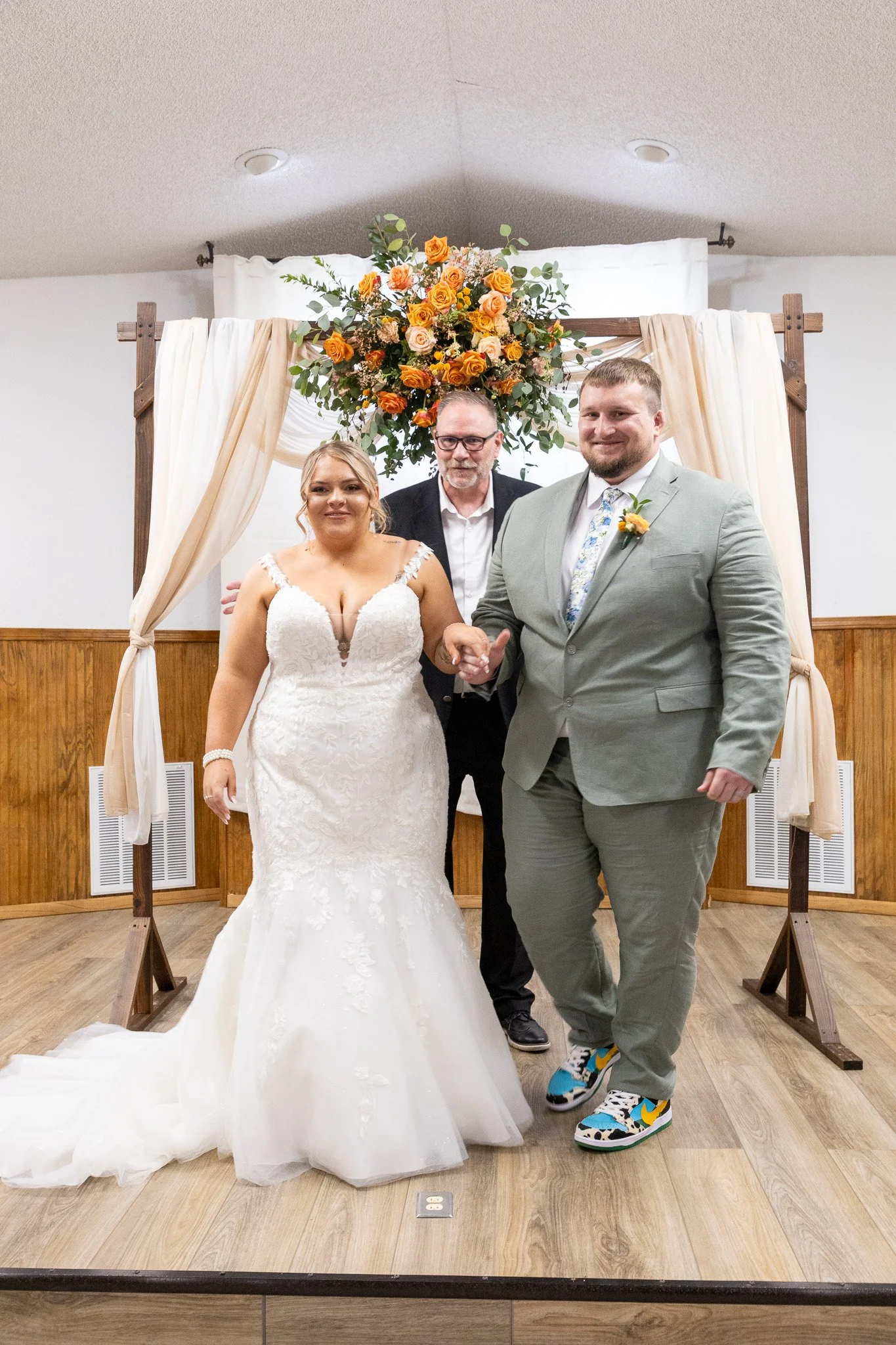 A newlywed couple standing in front of a decorated wedding arch, with a man, possibly the officiant, behind them. The bride is in a white wedding gown, and the groom is in a gray suit and colorful sneakers. The arch is adorned with orange and peach r