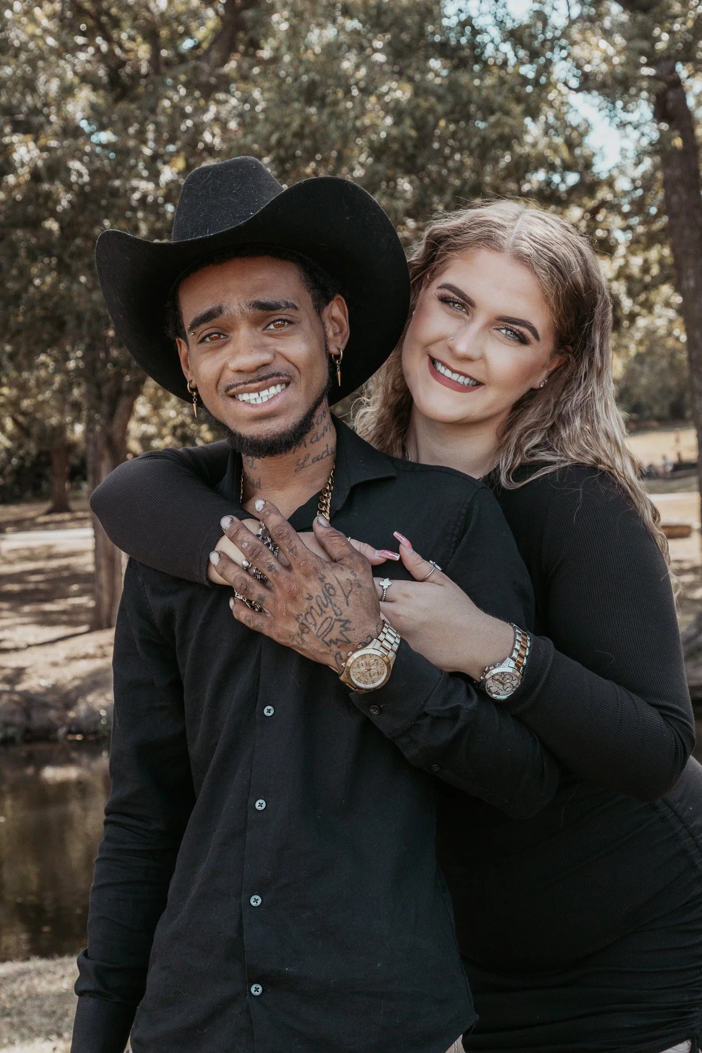 A smiling couple outdoors, the woman hugging the man from behind, both wearing black clothing. The man wears a black cowboy hat, has tattoos, jewelry, and a gold watch. The woman has curly blonde hair and also wears jewelry, both smiling at the camer