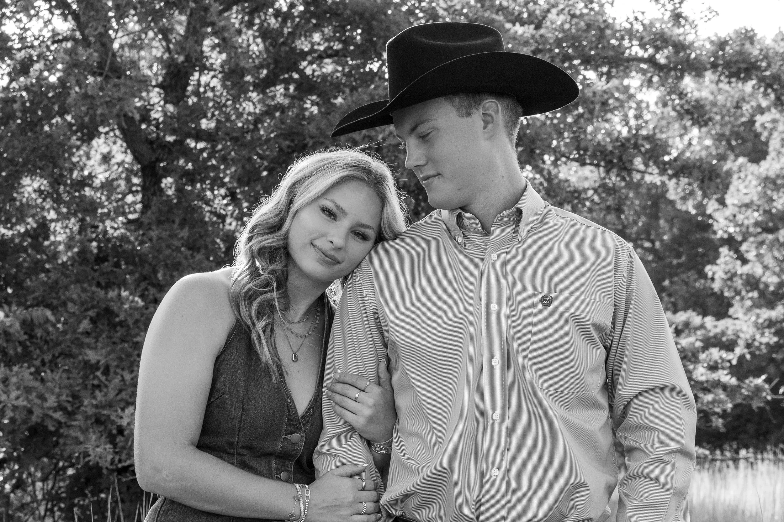 A black and white photo of a young couple outdoors with trees in the background. The woman is smiling, leaning her head on the man's shoulder, and wearing jewelry. The man is wearing a cowboy hat and looking down slightly, with his arm around the wom