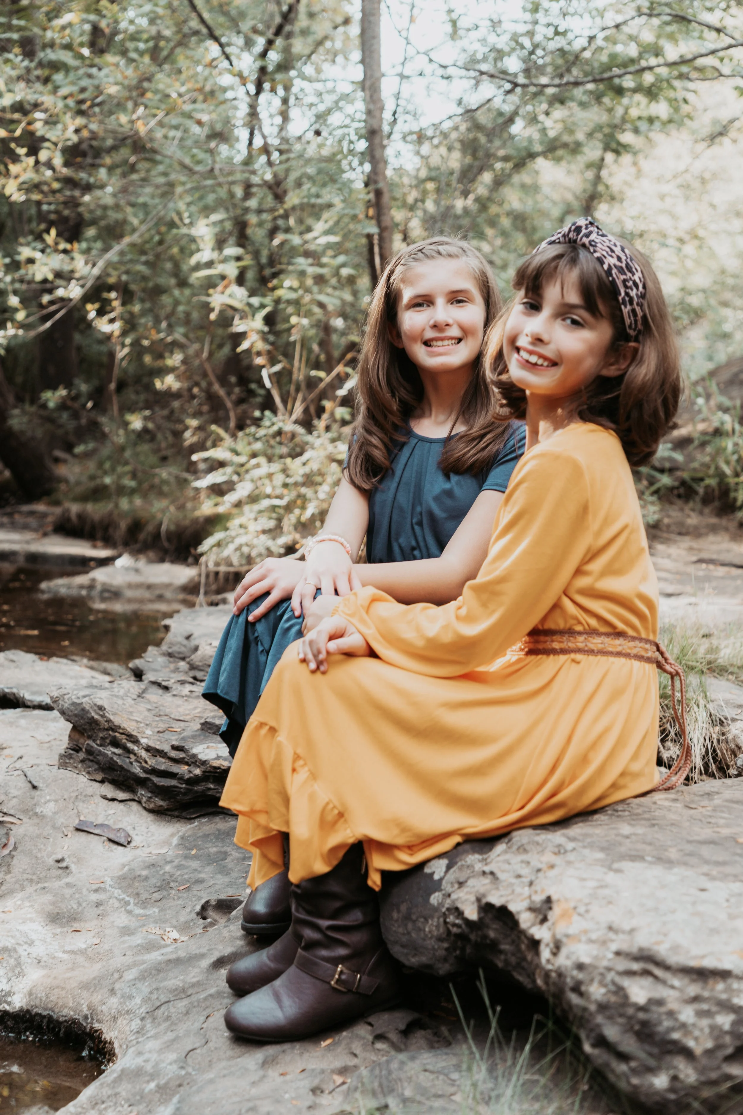 Two young girls sitting on a large rock near a creek in a wooded area, smiling at the camera.