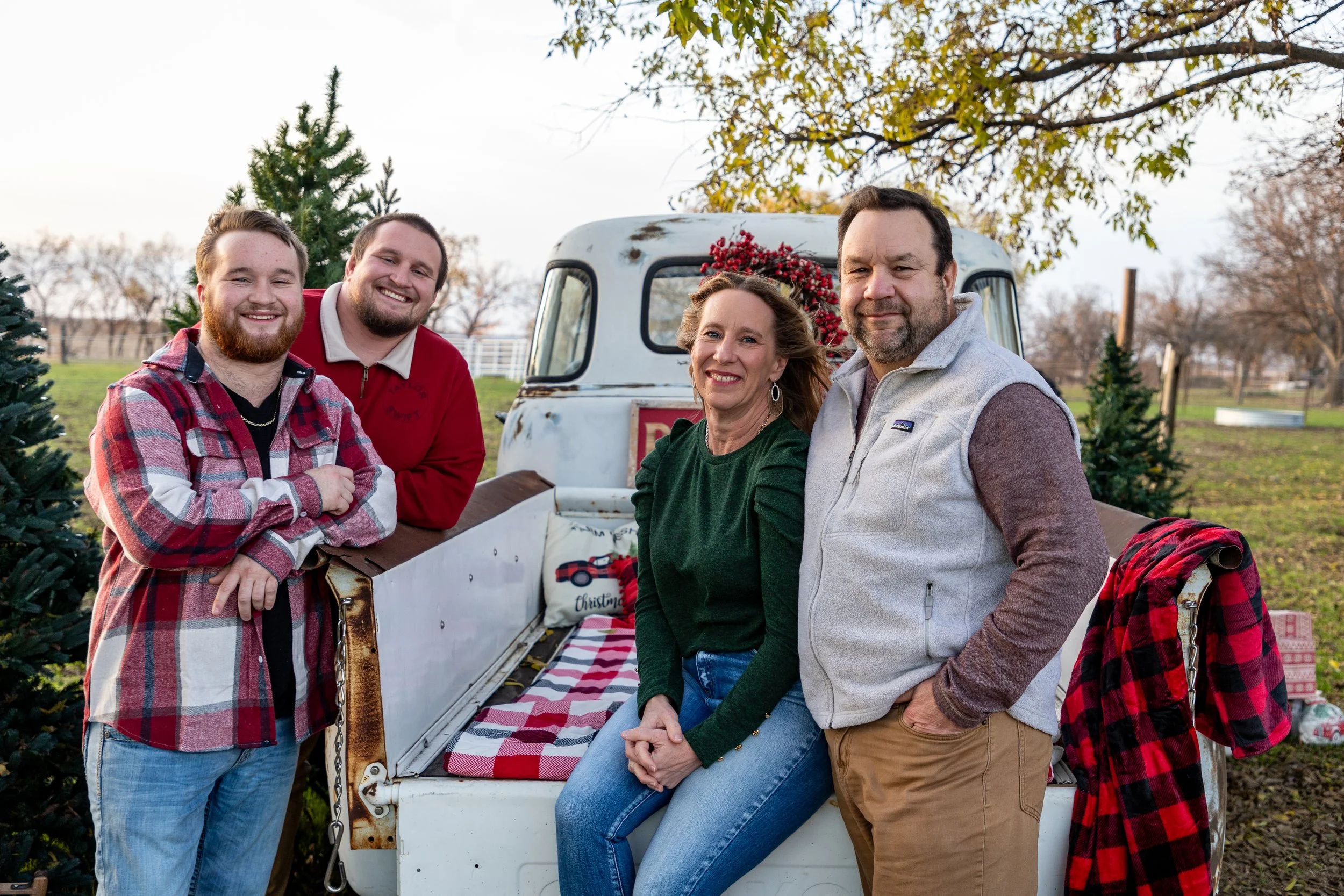 Family standing around the back of a vintage pickup truck decorated with Christmas wreaths and ornaments outdoors, with Christmas trees nearby and autumn trees in the background.