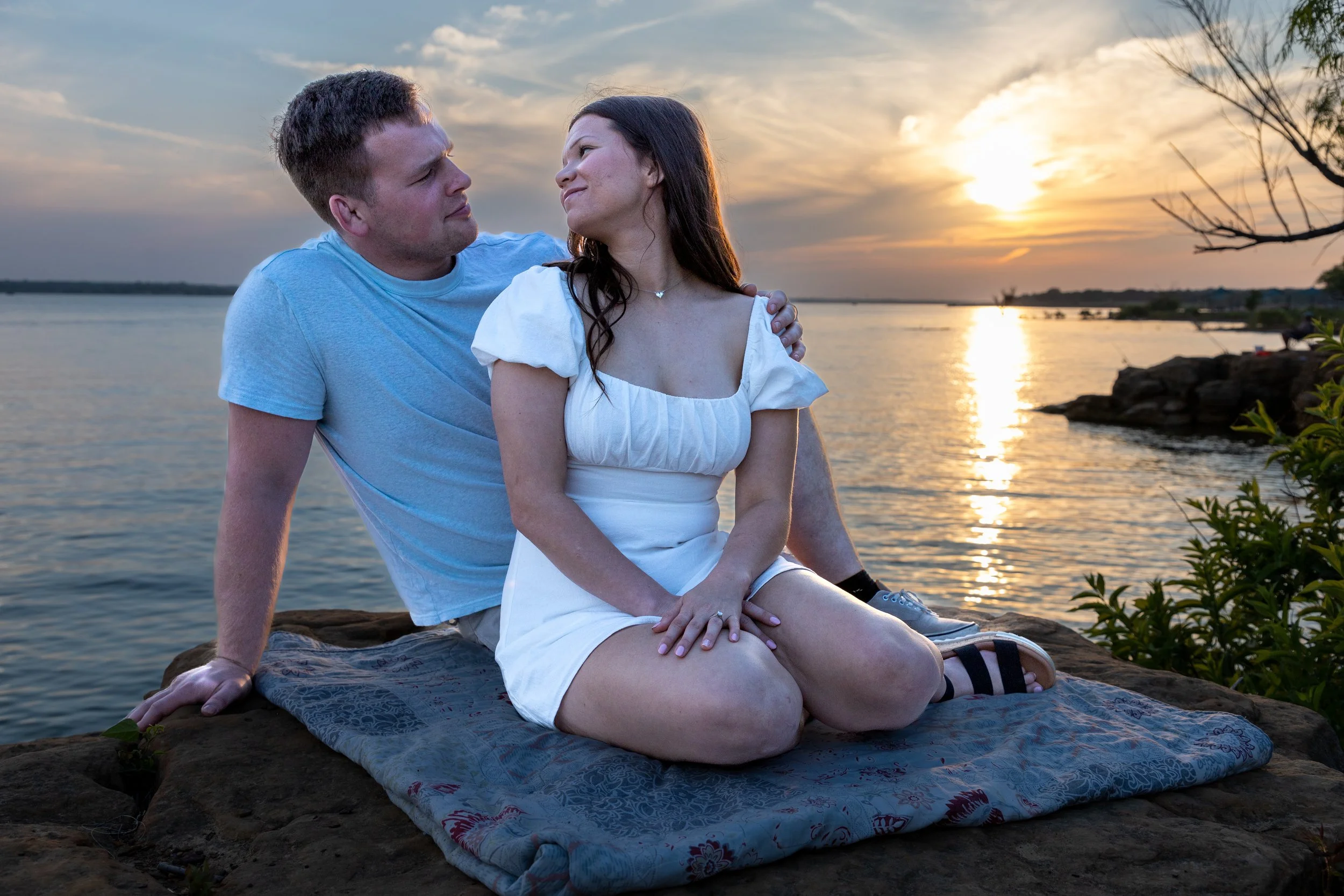 A couple sitting on a blanket by the water during sunset, gazing at each other