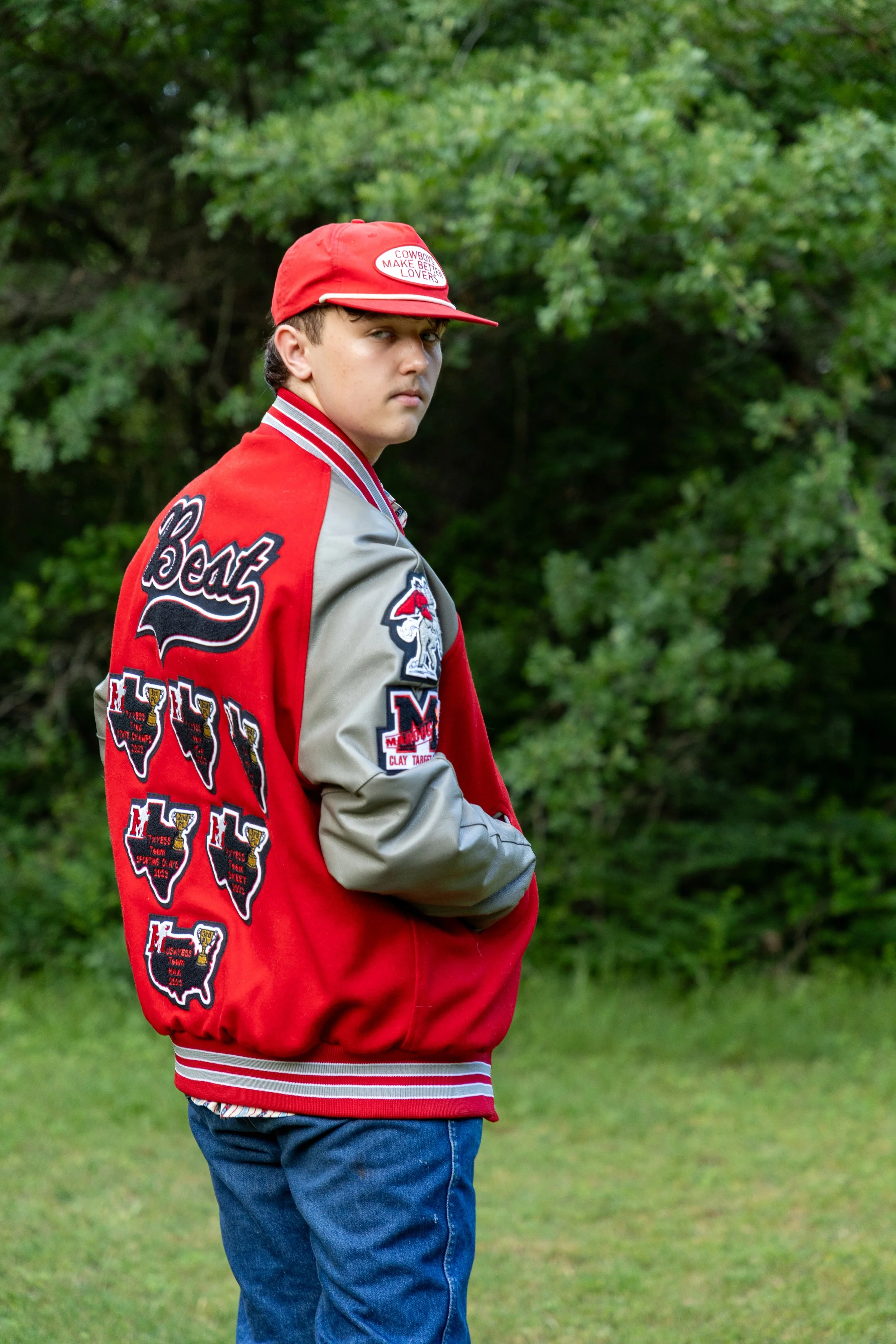 A teenage boy standing outdoors on a grassy area with trees in the background. He is wearing a red and gray varsity jacket with patches, a red cap with white text, and blue jeans, looking over his shoulder with a serious expression.