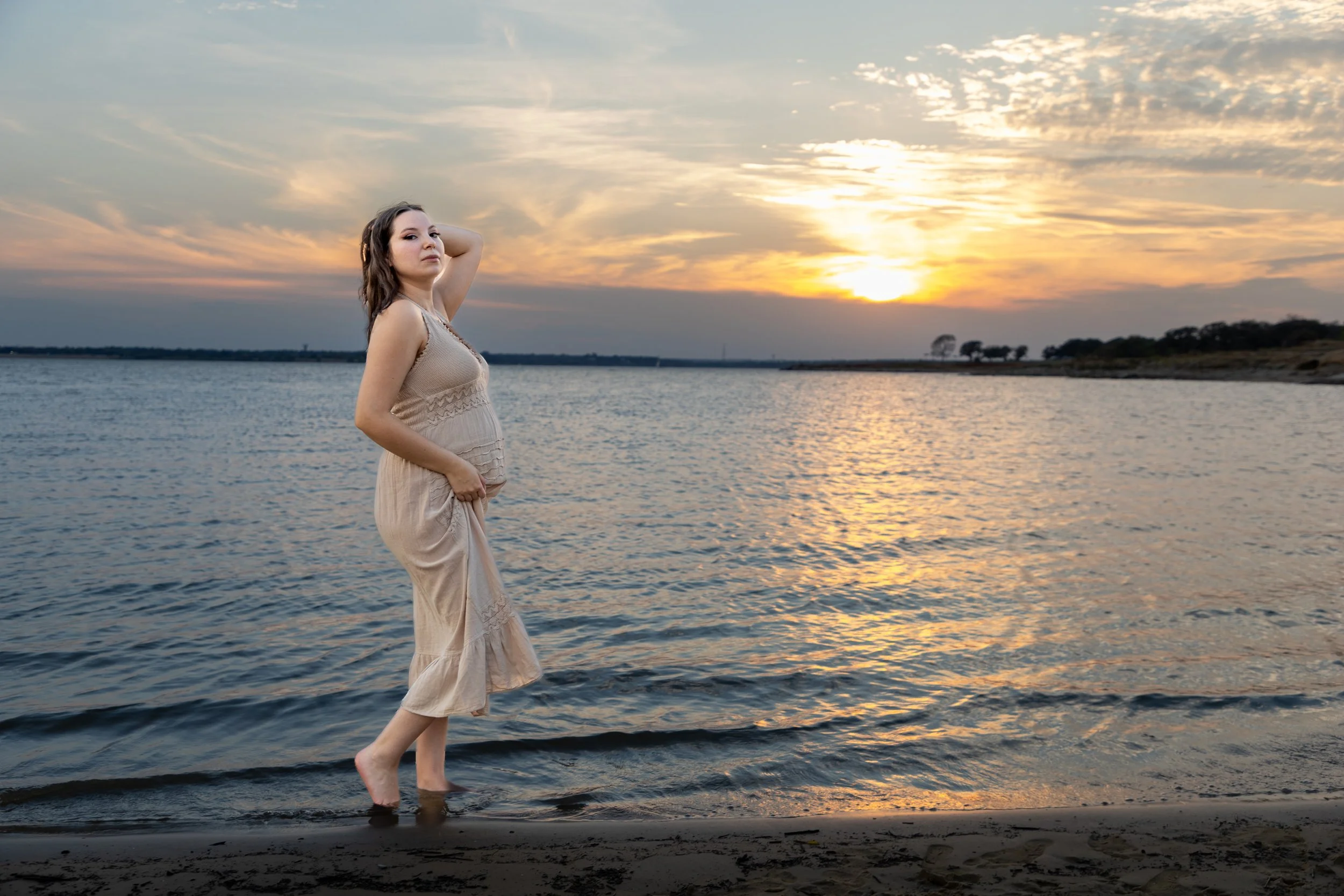 A woman in a beige dress standing in shallow water at sunset on a beach.