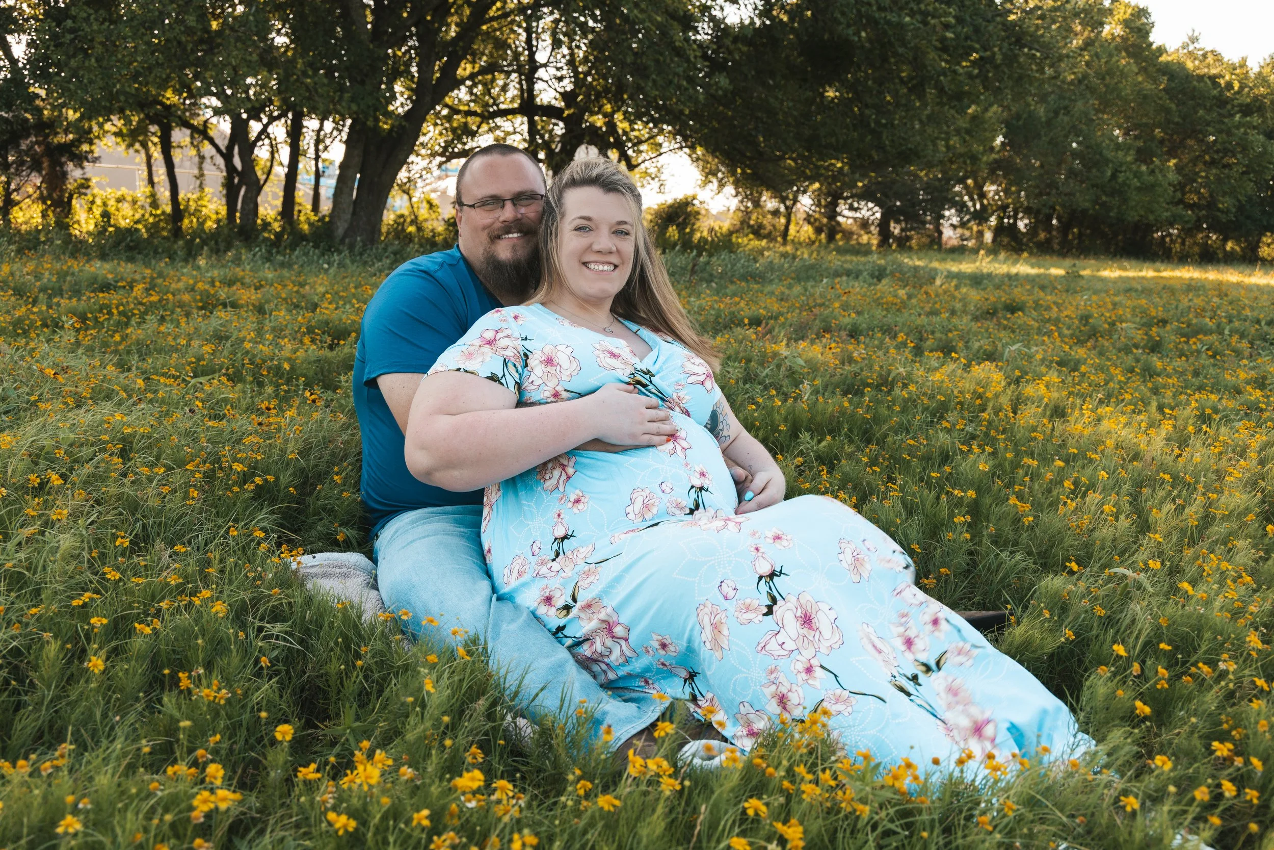 A smiling couple sitting on the grass in a field of yellow flowers with trees in the background during sunset.