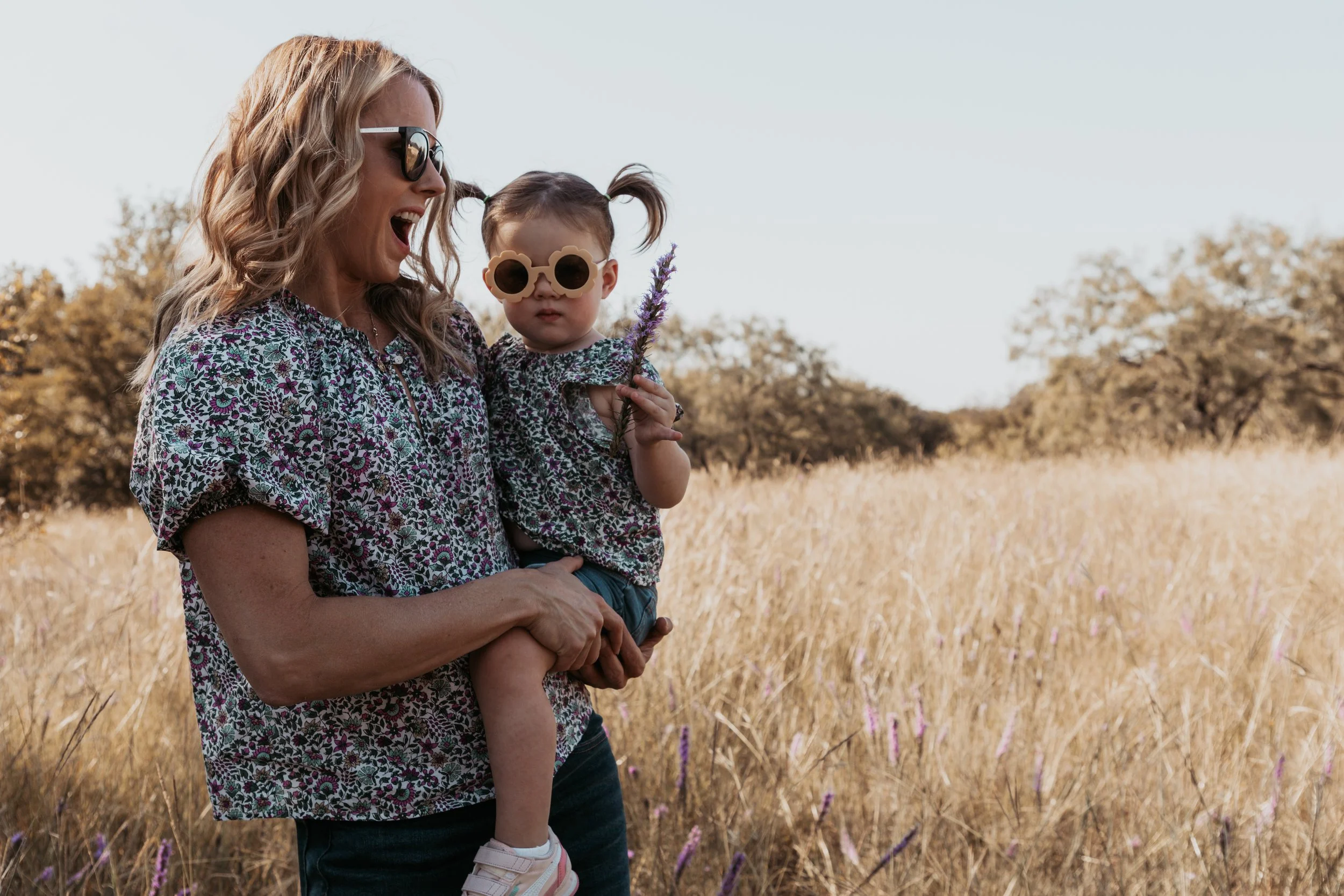 A woman holding a young girl in a grassy field with trees in the background. Both are wearing patterned shirts and sunglasses; the girl is holding a purple flower.