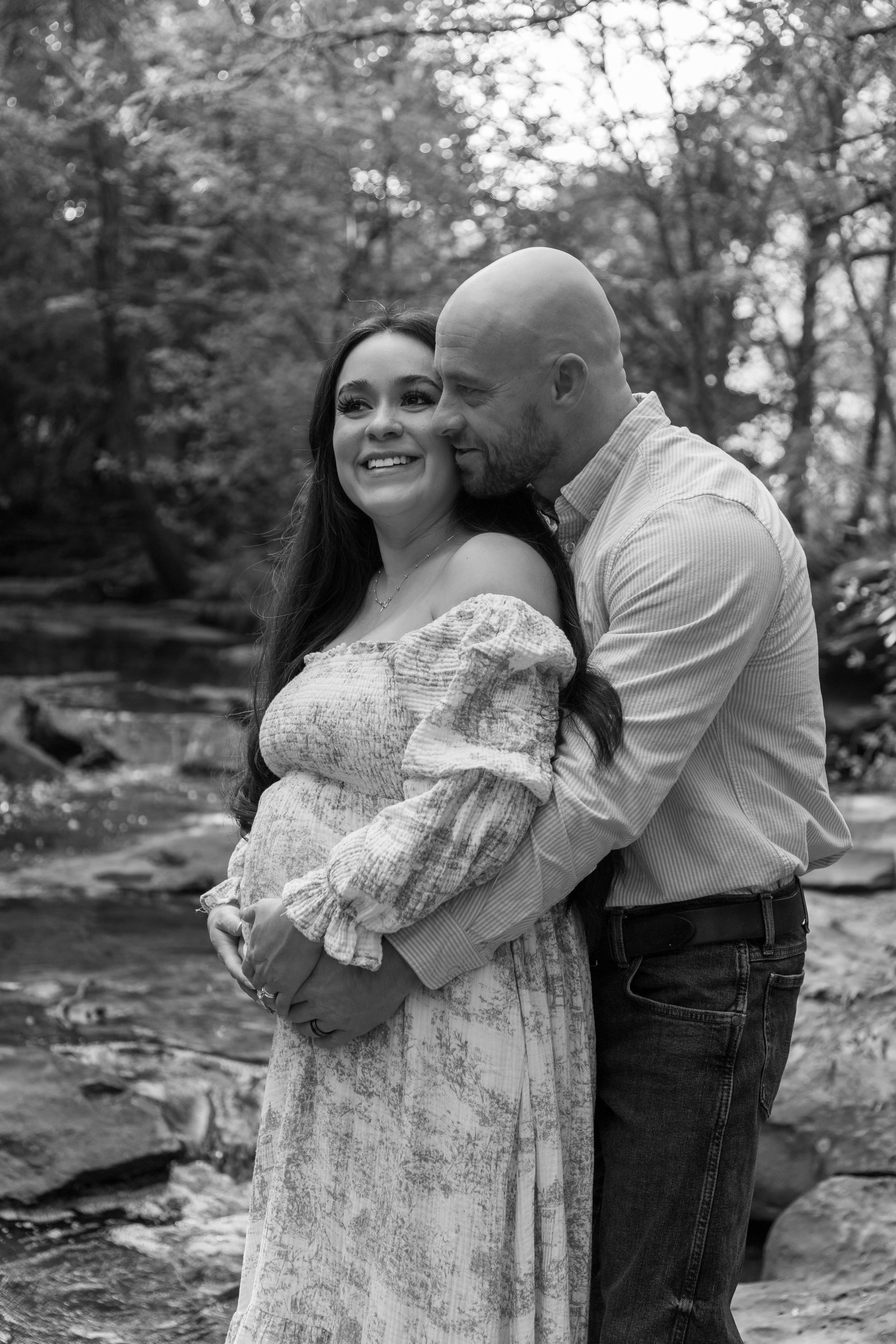A black and white photo of a smiling woman and a man embracing near a stream in a wooded area.