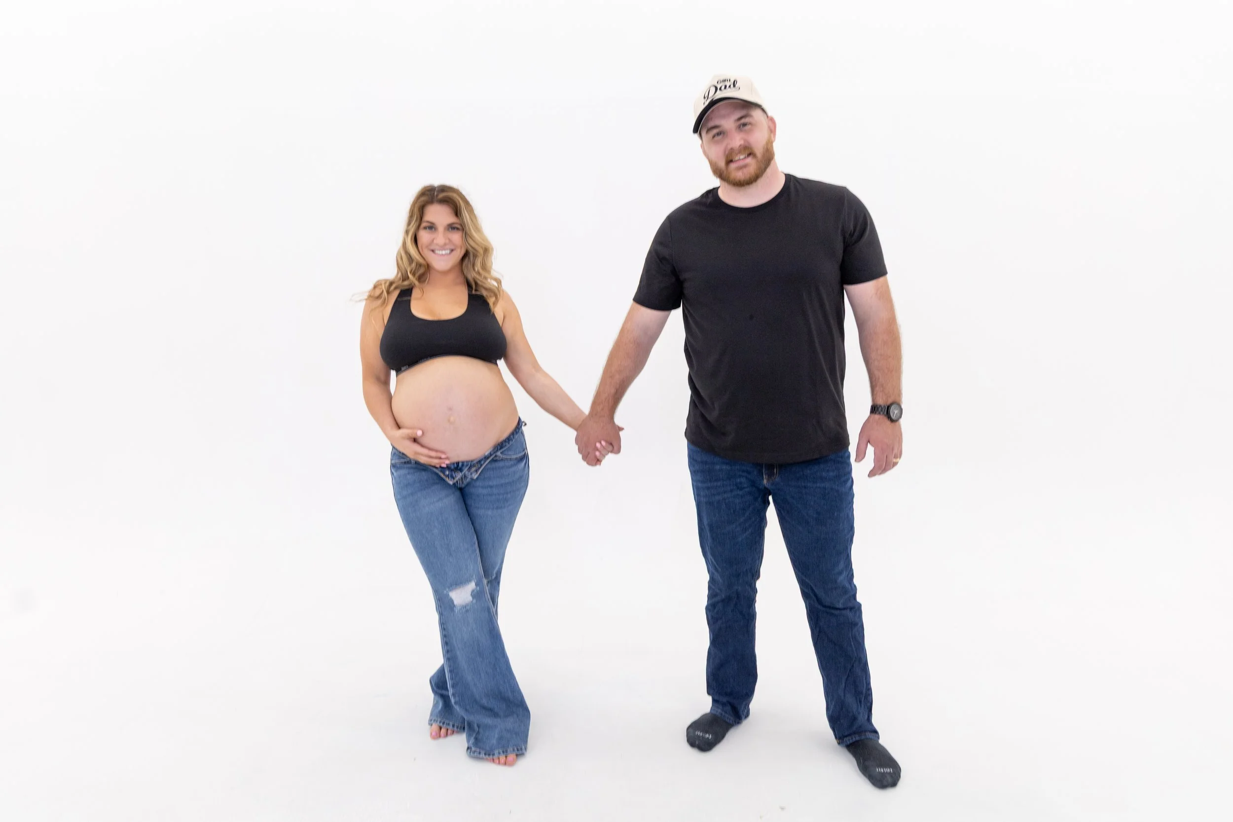 A smiling pregnant woman holding her belly and a man holding her hand, both standing against a plain white background.