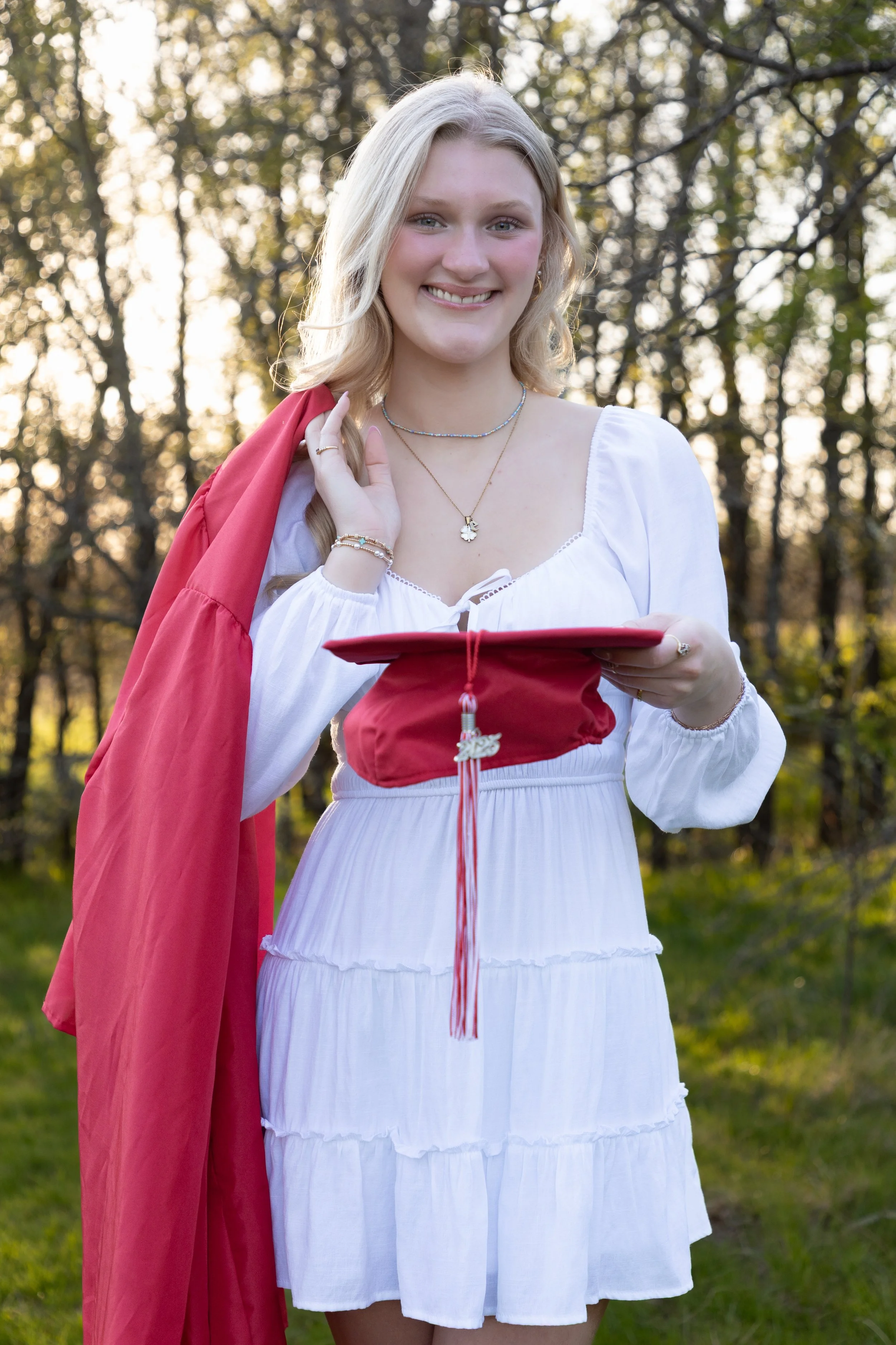A young woman in a white dress holding a red graduation cap with a tassel and a diploma, standing outdoors among trees, smiling.