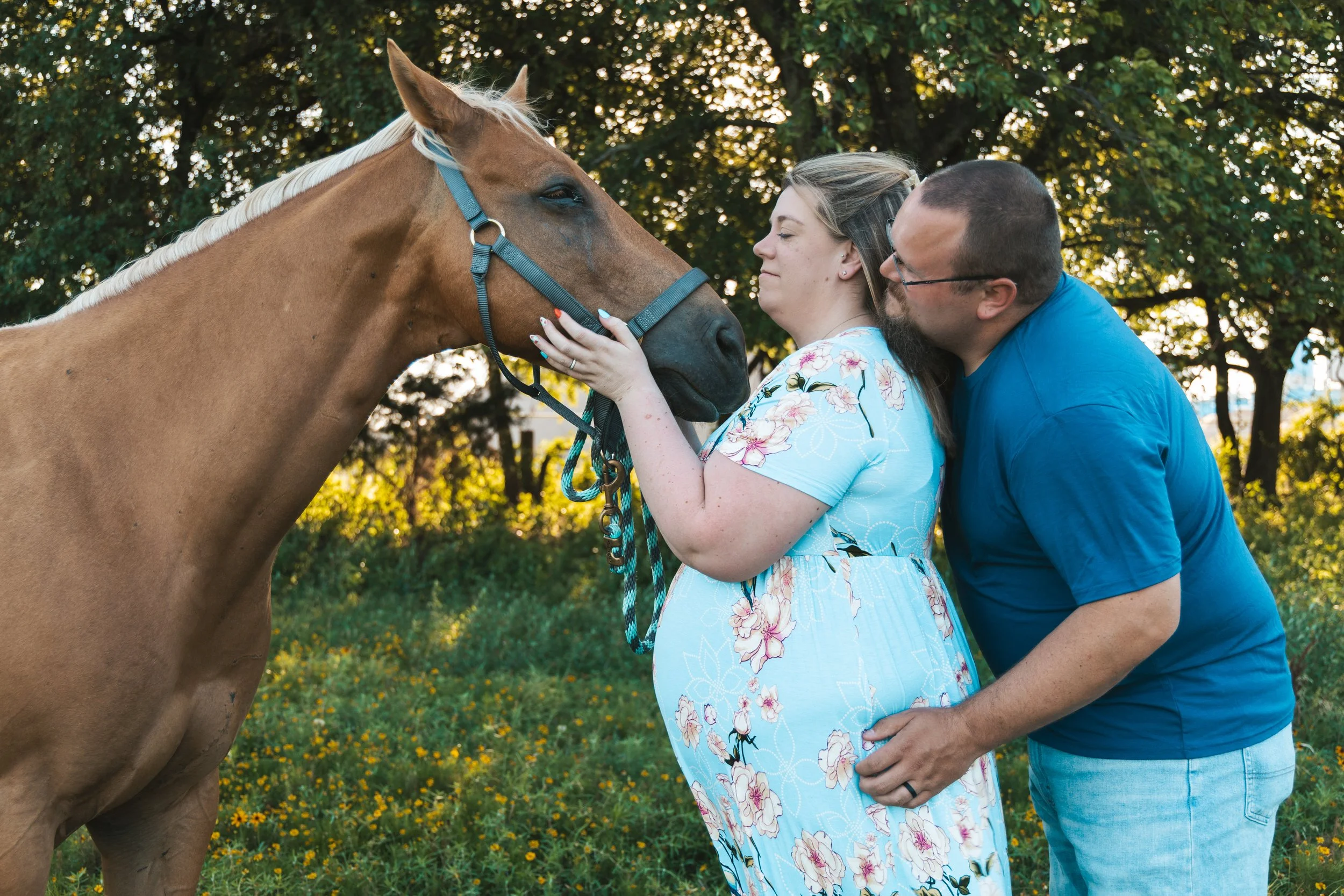 A woman and a man are standing outdoors, touching foreheads with a brown horse, under trees with sunlight filtering through the leaves.