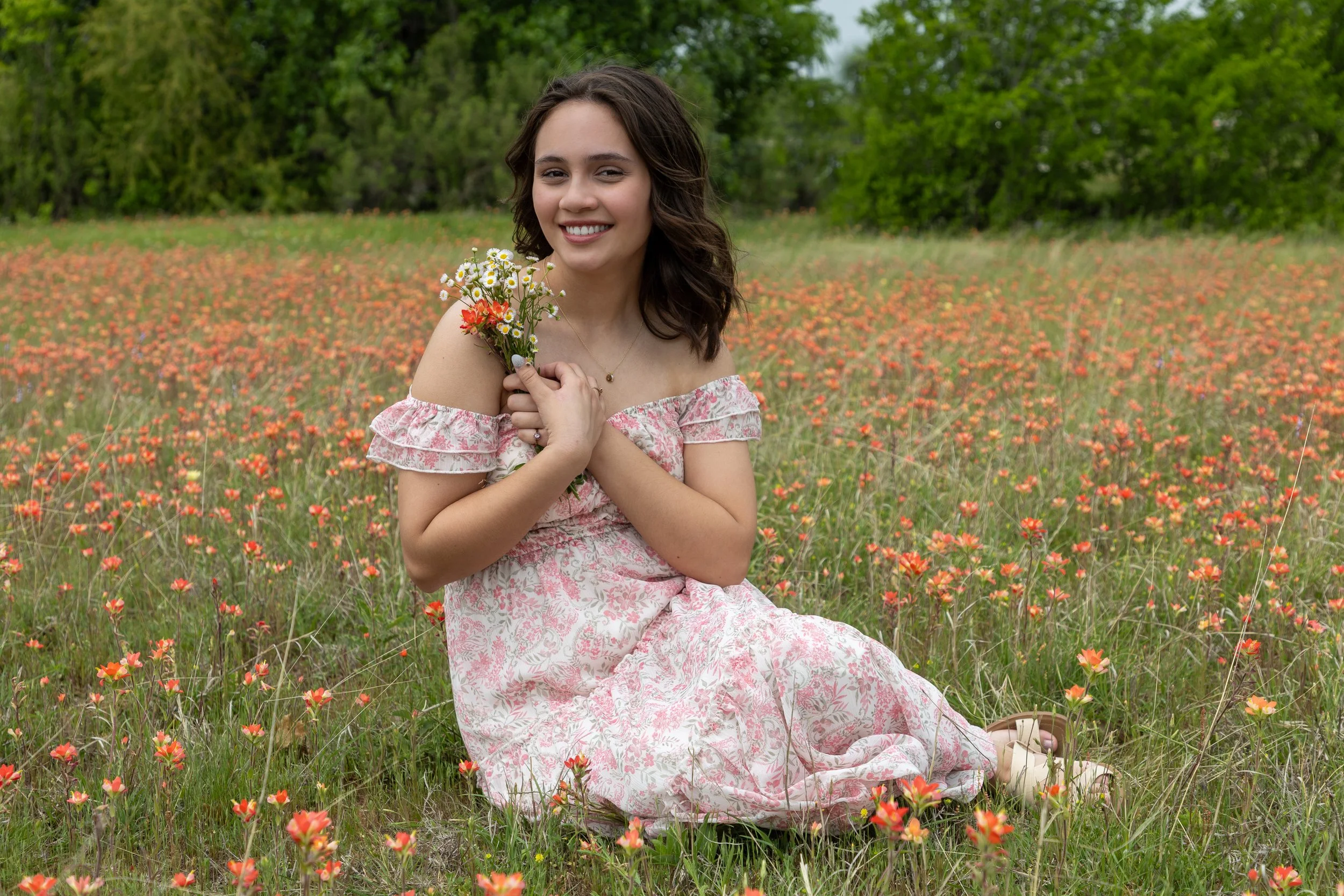 A young woman sitting on a grassy field filled with small orange flowers, holding a bouquet of wildflowers, and smiling. She has wavy brown hair and is wearing a pink floral off-the-shoulder dress. The background features green trees and a cloudy sky