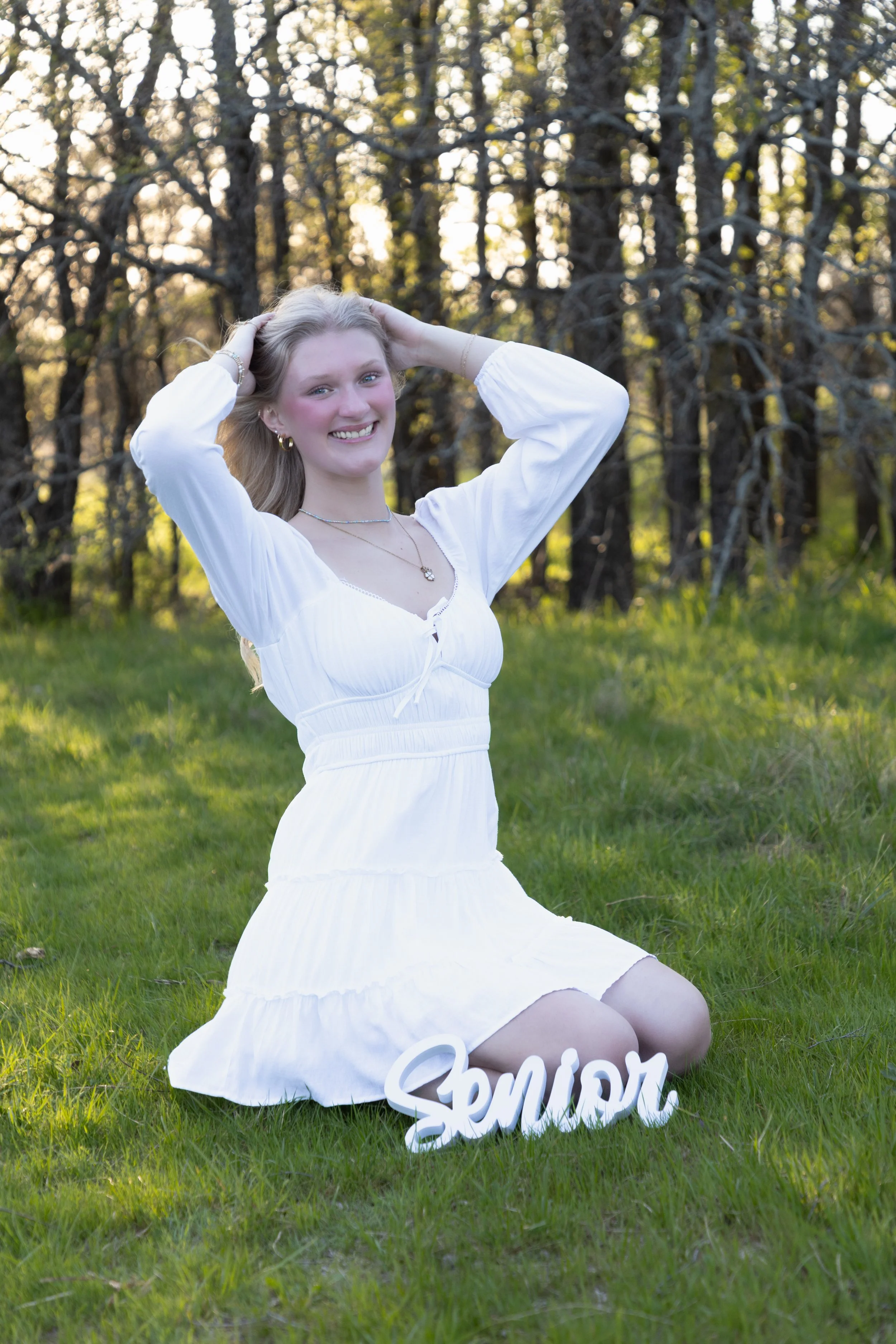 A smiling young woman in a white dress kneels on green grass in a park-like setting with trees and sunlight in the background. There is a white decorative sign that reads "Senior" placed near her knees.